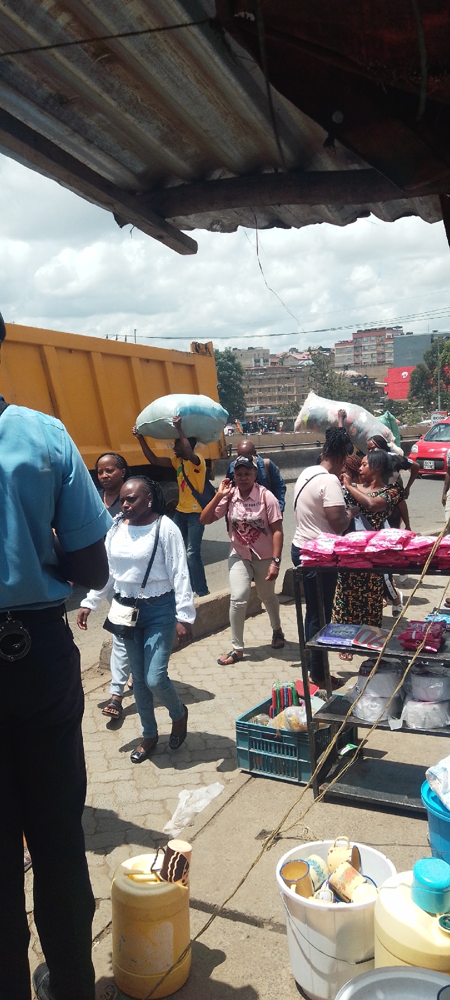 The image shows a busy street scene in Nairobi, Kenya. A yellow dump truck is parked near a sidewalk where pedestrians are walking. A man carries a large, light-colored bag over his head. A woman in a white blouse and blue jeans walks towards the viewer. A vendor's stand displays goods for sale. In the foreground, there are buckets and containers with various items. A man in a blue uniform is seen on the left side of the frame. Buildings are visible in the background.