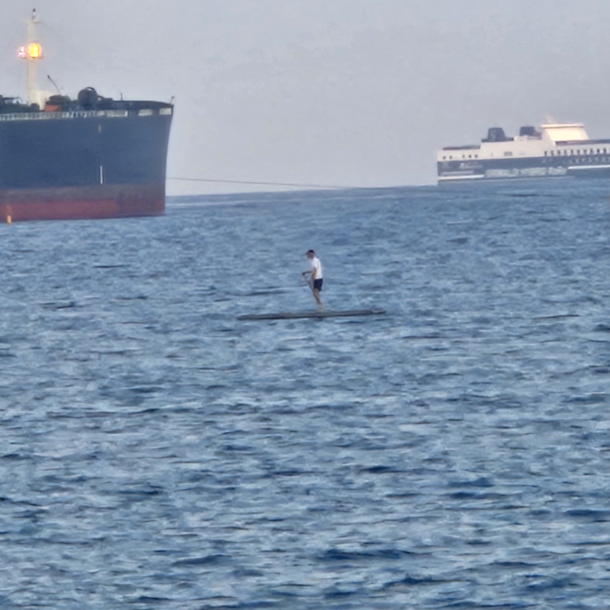 In the Savona harbor, a person is paddleboarding on choppy blue water. The person is wearing a white shirt and dark shorts and is holding a paddle. A large cargo ship is docked in the background on the left, and a ferry is visible further out on the right. The sky is overcast.