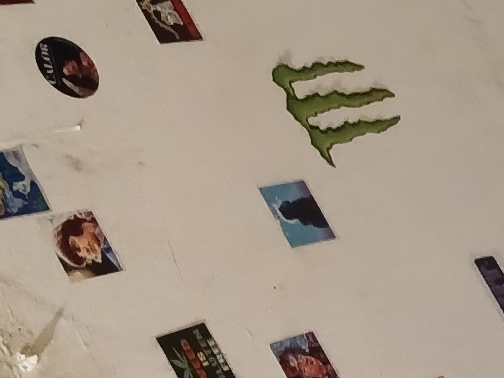 The image displays a light-colored, possibly off-white wall or ceiling surface adorned with a collection of stickers and small posters, suggesting a casual, personalized space. Dominating the upper right quadrant is a prominent, textured green Monster Energy logo, characterized by its three distinctive claw-mark stripes.
Several other stickers are visible across the surface. In the upper left, a circular black sticker features the word "CALOR" in white text, along with a partially visible face or graphic in red tones. Below this, a small rectangular sticker shows an abstract or ornate design in shades of blue and white. Further down and to the left, another rectangular sticker displays a complex pattern in dark red, orange, and brown hues, possibly depicting a face or skull.
In the lower left, a distinct rectangular sticker features a green cannabis leaf against a dark background. Accompanying text includes "WEED IS SEED," "PLANTEN," "FREE," and "IS REAL," though some words are partially obscured. To the right of this, another rectangular sticker shows a person's profile or head rendered in dark red and brown. Another small, rectangular sticker in blue and black depicting an abstract shape is visible in the middle right of the frame.
The overall impression is that of a wall or ceiling decorated with various decals and small artworks, reflecting diverse interests. The lighting is even, and no specific time of day or weather conditions are discernible. Based on the provided context, this scene is located in Buenos Aires, Argentina.