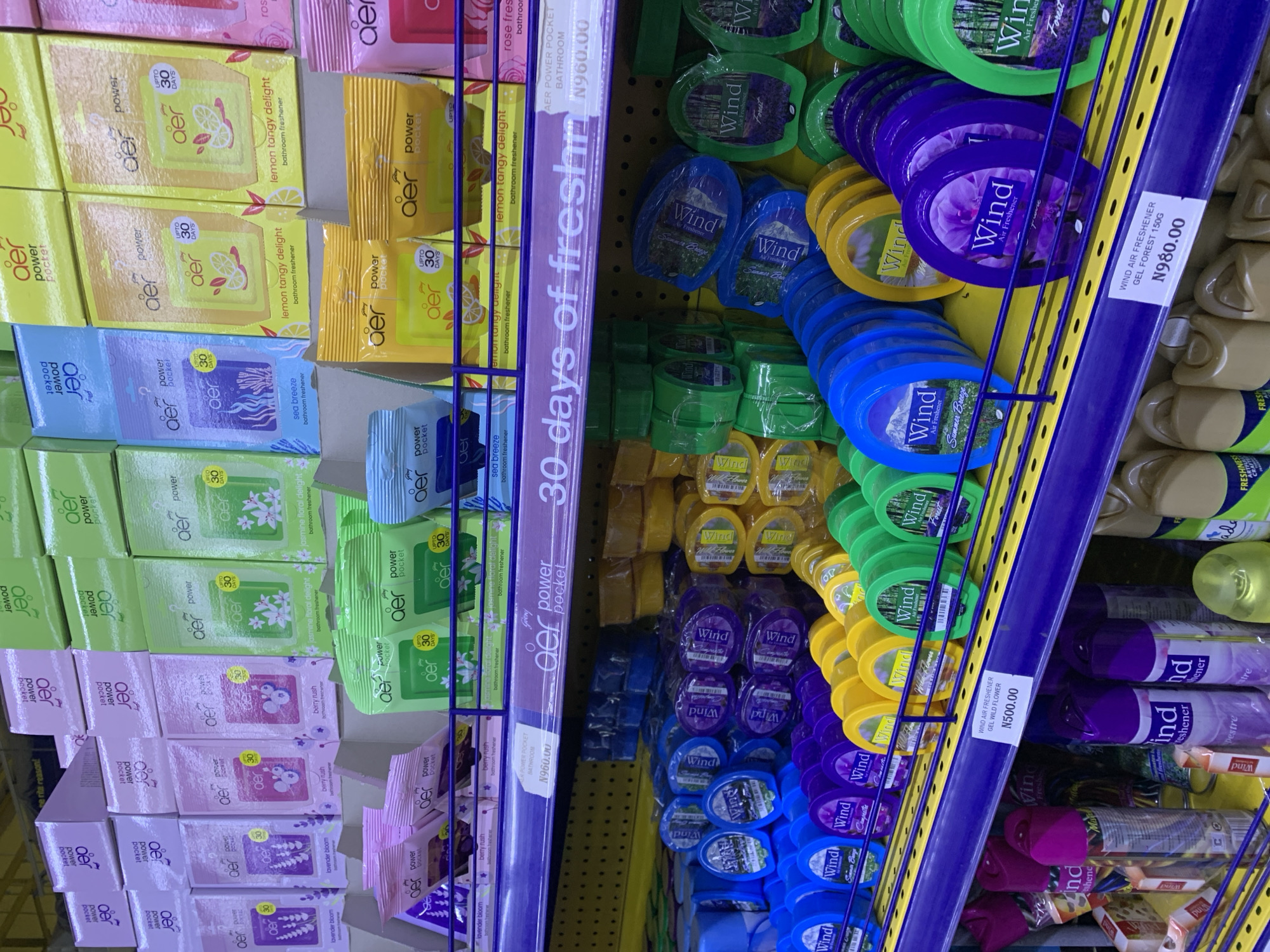 The image shows a close-up view of shelves stocked with various air fresheners in a retail setting. The products are primarily "Aer" brand pocket air fresheners in different scents like "Lemon Tangy Delight," "Jasmine Floral Delight," "Sea Breeze," "Berry Rush," and "Lavender Bloom." These are packaged in small, colorful sachets, predominantly in yellow, green, pink, and purple.
Alongside the "Aer" products, there are "Wind" brand air fresheners displayed. These include gel air fresheners in round containers with lids, also in various colors like green, yellow, blue, and purple, suggesting different fragrances such as "Forest" and "Wild Flower." Some of these "Wind" air fresheners have labels indicating their scent and price.
The shelves are made of purple and yellow metal. A prominent vertical strip on the left side of the image has text that reads "30 days of freshn" and a price of "N960.00". Another price tag visible on the right reads "N500.00" for "WIND AIR FRESHENER GEL WILD FLOWER" and another for "WIND AIR FRESHENER GEL FOREST 150G N980.00". The overall impression is of a well-organized display of household air freshening products in a store. There are no people visible in the image. The lighting suggests an indoor environment, possibly a supermarket or a dedicated home fragrance store.