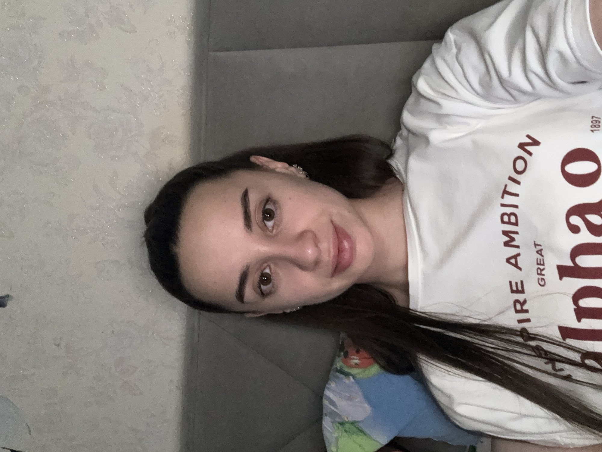 A young woman is depicted in a close-up portrait, looking directly at the camera with a gentle smile. She has long, dark hair, neatly styled eyebrows, and brown eyes. She is wearing a white t-shirt with text printed on it. Visible text on her shirt reads "EMPIRE AMBITION GREAT 1897", and below this, in larger red letters, "alphao", where the final 'o' is notably larger and styled. A small, sparkly earring is visible on her right ear.
She appears to be reclining or lying down, with a gray couch or headboard behind her. To her left, a light-colored wall with a subtle floral or damask pattern is visible. Part of a colorful pillow or blanket with abstract blue, green, and orange patterns is seen in the lower portion of the image. The setting is indoors, with soft, even lighting, suggesting a casual environment in Goncharka, Russia.
