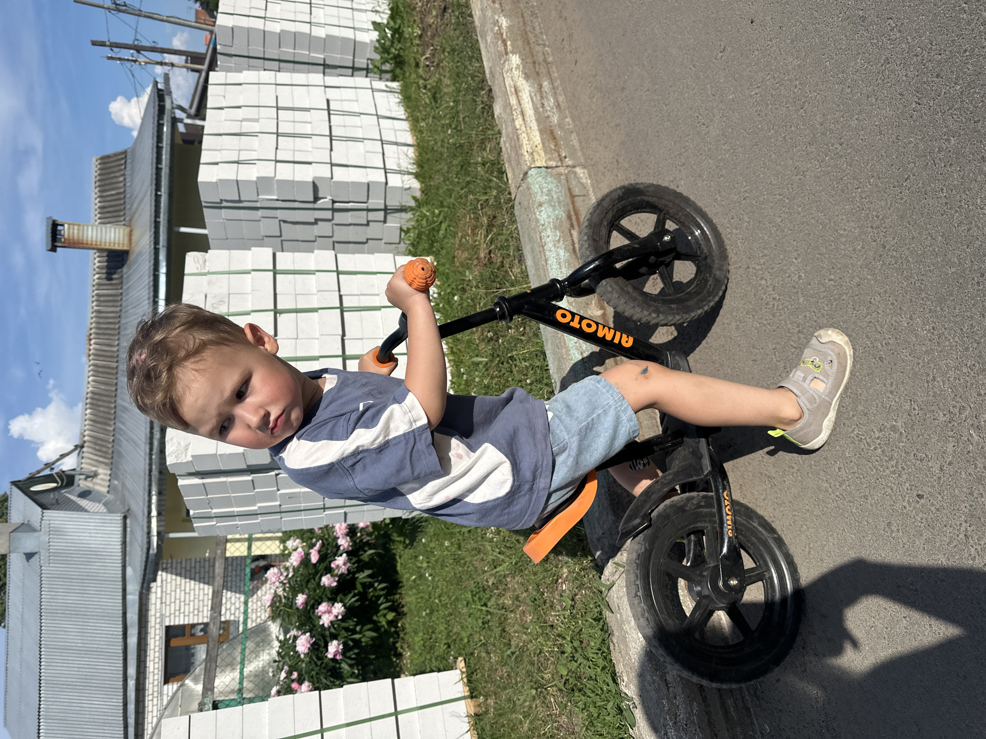 A young Caucasian boy with light brown hair is sitting on a black balance bike with orange handlebars and seat. He is wearing a blue and white t-shirt and light denim shorts. The boy is looking to his left with a thoughtful expression. The balance bike has "AIMOTO" written in orange on the frame. He is on an asphalt path next to a curb. Behind him, there are stacks of white building blocks, a bush with pink flowers, and a building with a grey, corrugated metal roof and yellow siding. The sky is blue with some white clouds. The scene appears to be outdoors in a residential or construction area.