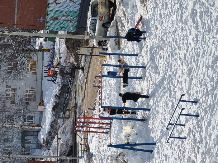 The image presents an outdoor winter scene in Chaikovsky, Russia, dominated by extensive snow cover under clear skies. In the foreground, a blue and red multi-station calisthenics rig occupies a snow-covered area. Three males are present around this equipment. One male, dressed in a light grey short-sleeved top and dark pants, is performing a horizontal suspension exercise (human flag) on the blue bars. Another male, wearing a black shirt and dark pants, stands near the blue uprights, resting his left hand on a bar. A third male, in dark clothing, is partially visible further left on the structure. A black jacket or similar garment is draped over a red bar section to the right.
In the mid-ground, a paved road runs horizontally across the frame, with a silver Hyundai Santa Fe SUV (license plate X999YE159 RUS) parked on its right side, facing left. Adjacent to the vehicle, a brick building features a prominent mural with stylized white figures on a teal and green background. A utility pole stands near the vehicle. In the background, a light-colored multi-story residential building with numerous windows rises behind a snow-covered, fenced-off playground containing colorful slides, swings, and a small basketball hoop. Large snow accumulation is evident across all ground surfaces.