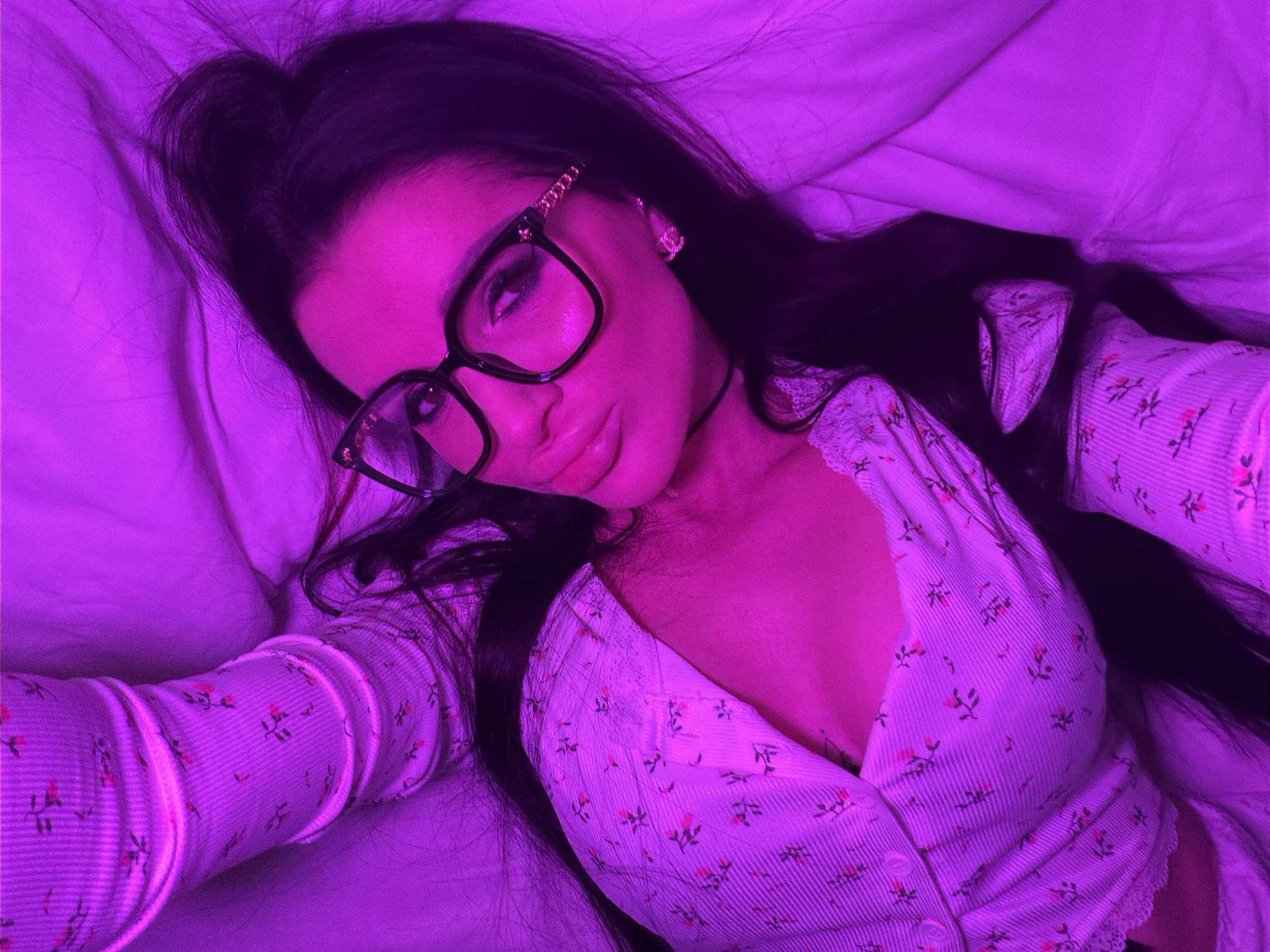 A young woman with dark hair pulled back into a high ponytail lies on a white bed in a room illuminated by purple light. She is wearing large, square-framed glasses with ornate detailing on the temples. She wears a light-colored, floral-patterned long-sleeved shirt with a V-neck and buttons. The environment is a bedroom setting with bedding visible. There is no information available to determine if this image was taken in Voronezh, Russia.
