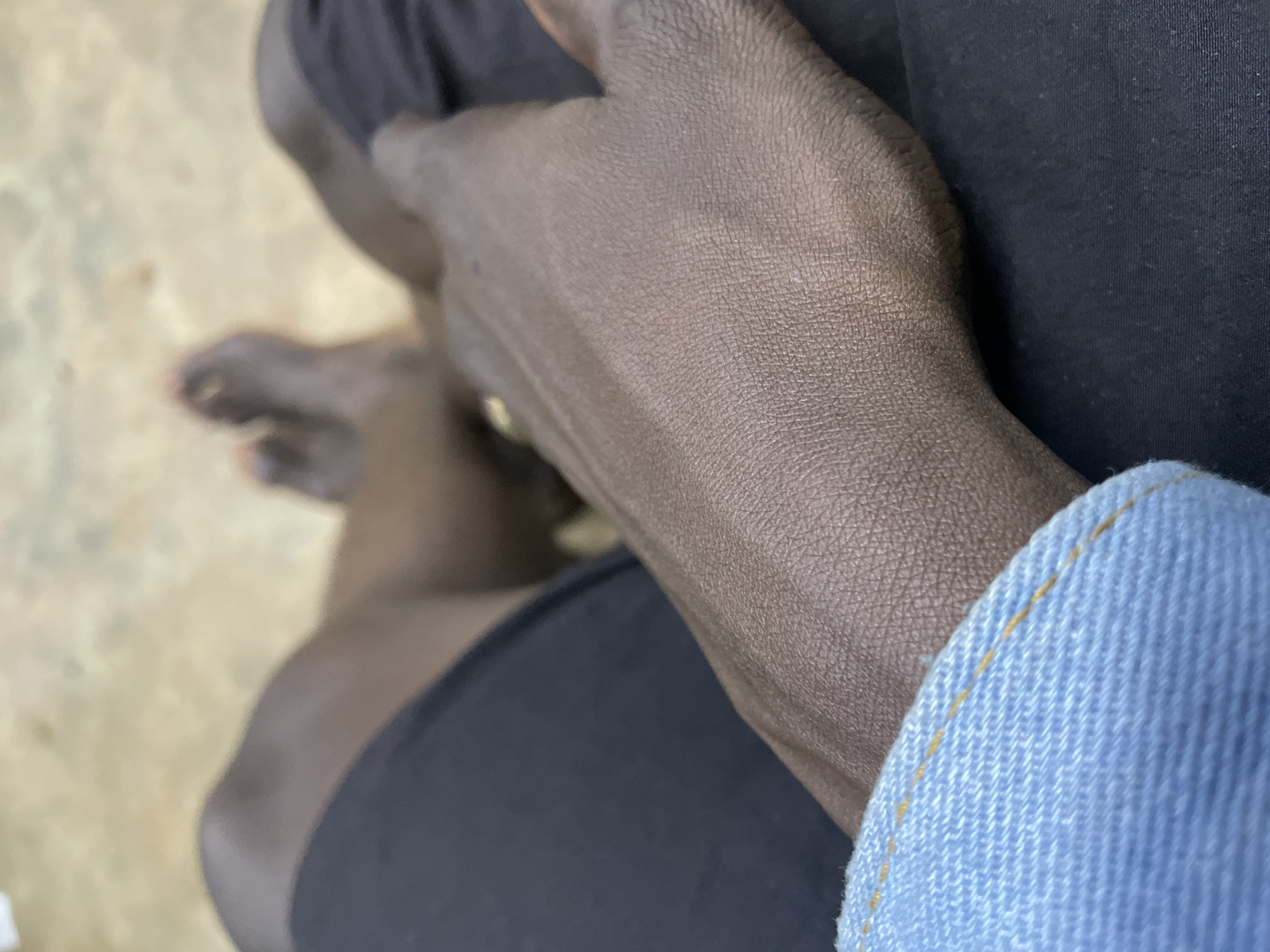 The image shows a close-up view of a person's hands and feet, resting on dark fabric. The person's skin is dark brown. The hands are positioned on the lap, with one hand resting on top of the other. The feet are visible in the background, with toes slightly spread. A light blue denim cuff is visible in the foreground, suggesting the person is wearing a denim shirt or jacket. The background is blurred and appears to be an outdoor setting with a light-colored, textured surface, possibly concrete or pavement. There is no indication of the specific location within Durumi, Nigeria.