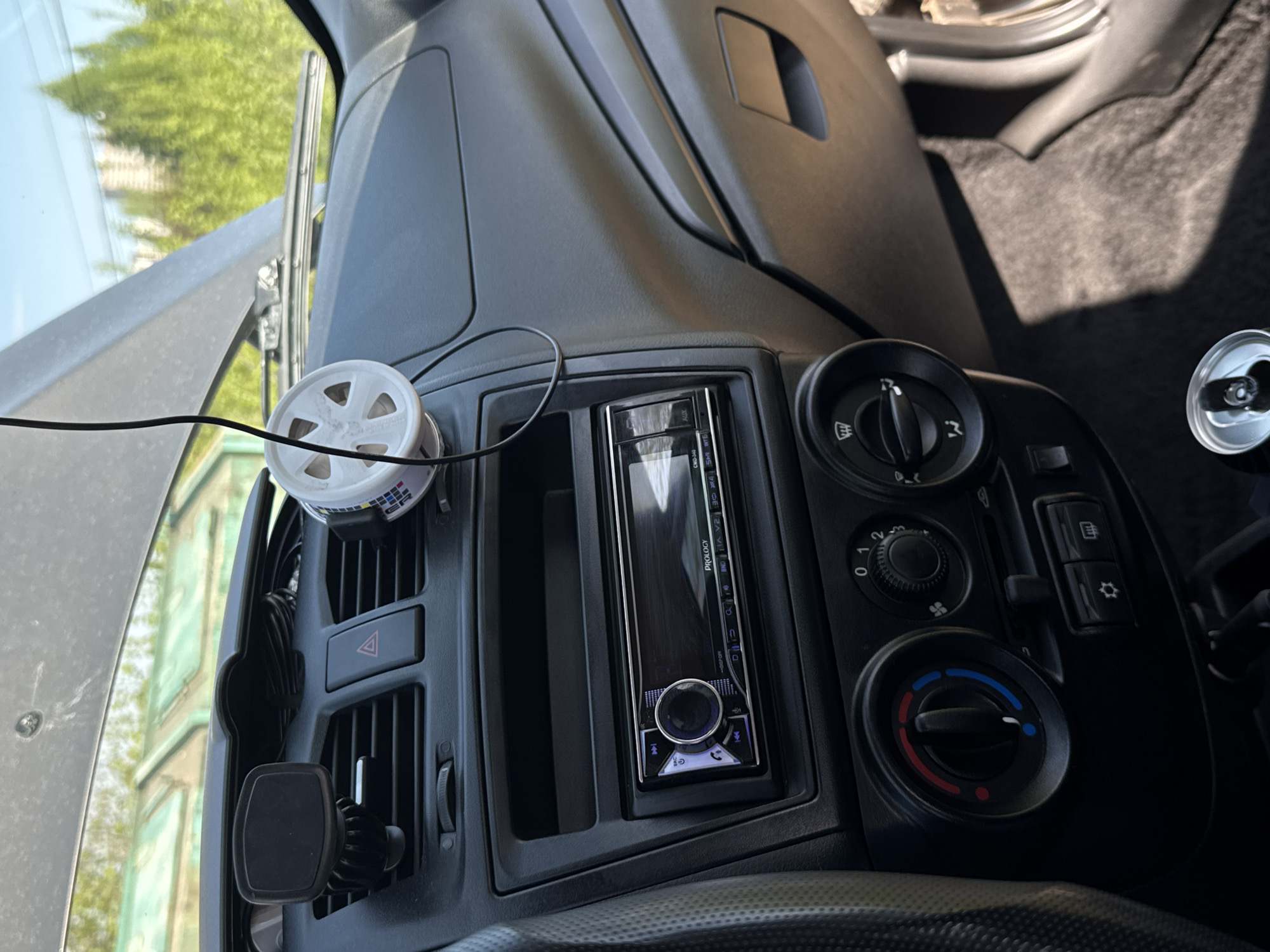 This is a close-up shot of the dashboard area of a vehicle, focusing on the center console. The console features a car stereo head unit, labeled "PROLOGY CMD-340," with a display screen. To the left of the stereo, there's a set of air conditioning vents with a car air freshener clipped to the top and a magnetic phone mount attached below. To the right of the stereo, are a set of knobs for adjusting the fan speed and temperature. A silver opened can of soda is on the right in the background. The environment suggests the vehicle is parked or stationary. The city of Perm, Russia is not evident from this image.