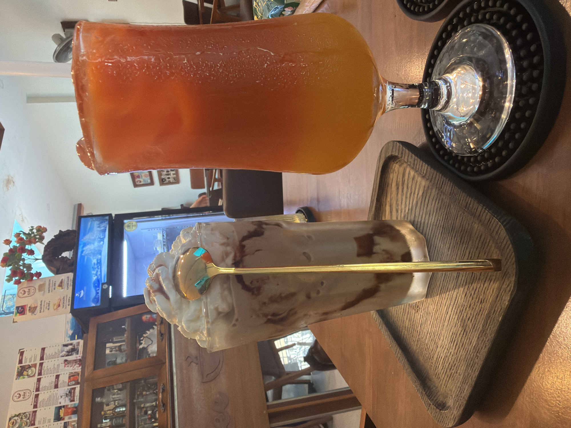A close-up shot shows two beverages on a wooden table in what appears to be a cafe in Bac Nha Trang Ward, Vietnam. On the left is a tall glass filled with a chocolate-colored beverage, topped with whipped cream and drizzled with chocolate sauce, and a golden spoon. The glass rests on a dark wooden tray. To the right, a stemmed glass contains a layered orange and yellow drink, possibly a juice or cocktail, with condensation on the glass. It sits on a black coaster. In the background, shelves filled with bottles and decorative items are visible, along with a refrigerated display case showing food items. A menu board is also visible on the wall. The lighting suggests an indoor setting.