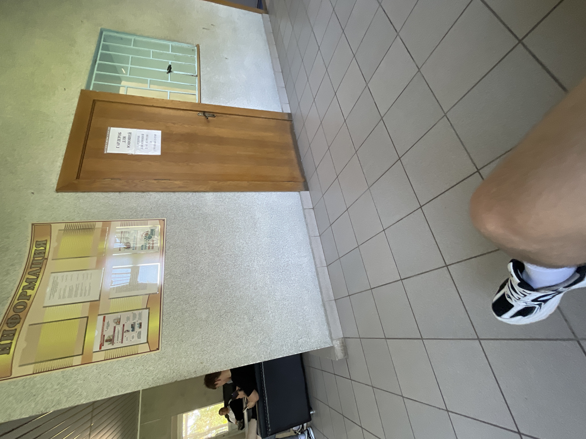 In a dimly lit interior hallway with tiled floors, an information board with Russian text hangs on a textured, light-colored wall. To the right of the board is a closed wooden door with a sign that reads "ГАРДЕРОБ ДЛЯ ЮНОШЕЙ" (Cloakroom for Boys). Next to the door, a barred blue window is visible. In the background, a person sits on a black sofa, looking at a phone. The foreground shows a person's leg and foot wearing a black and white sneaker and white socks. The scene appears to be inside a building, possibly an educational institution, given the presence of an information board and a cloakroom sign. While the exact location within Krasnye Baki is not specified, the context suggests it's a public or institutional space.