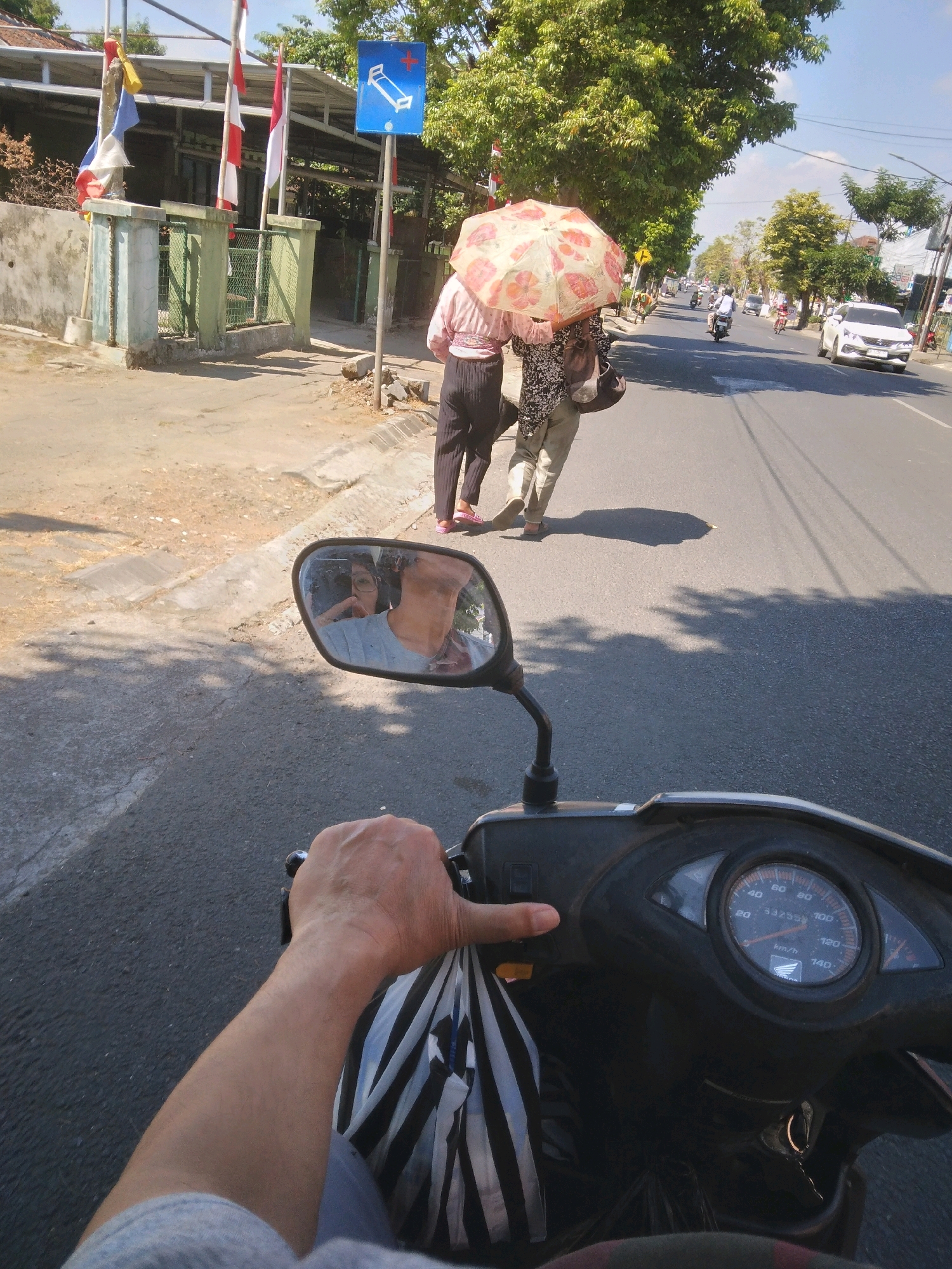 The image captures a street scene in Bancarkembar, Indonesia, from the perspective of someone riding a scooter. In the foreground, a hand is visible on the scooter's handlebar, with a striped bag hanging from it. The scooter's dashboard is also in view, showing a speedometer with a reading around 30 km/h.
On the sidewalk to the left of the road, two individuals are walking under a patterned umbrella, shielding themselves from the sun. One person is wearing a pink shirt and dark pants, while the other is dressed in a floral shirt and light-colored pants. They are casting long shadows on the ground. A street sign with a blue background and a white symbol of a bed and a plus sign is also present on the sidewalk.
Further down the street, other vehicles, including motorcycles and a white car, are visible, indicating active traffic. The environment appears to be a sunny day with trees lining the sides of the road. Buildings and fences are also discernible in the background. The reflection in the scooter's rearview mirror shows the rider holding the camera and wearing a grey t-shirt.