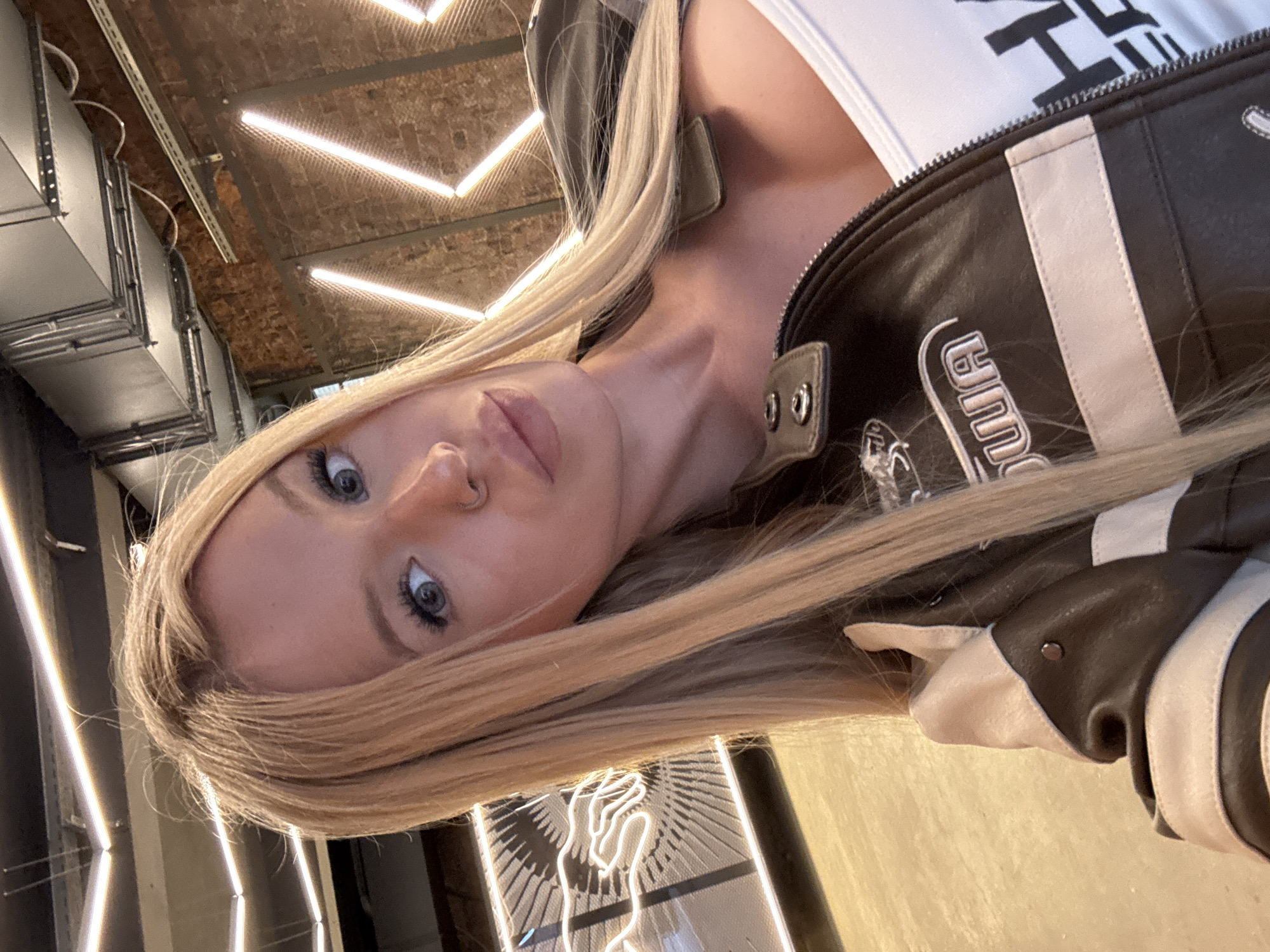 A young, blonde woman with blue eyes and a nose ring takes a selfie in what appears to be an indoor establishment. She is wearing a white tank top and a brown and beige leather jacket. The background features brick walls and industrial elements, with illuminated neon lights arranged in a geometric pattern. A neon sign depicting a hand is visible on the left side of the frame. The setting suggests a modern, trendy urban location, possibly a bar or restaurant. The specific location within Saint Petersburg, Russia, cannot be determined from the visual information provided.