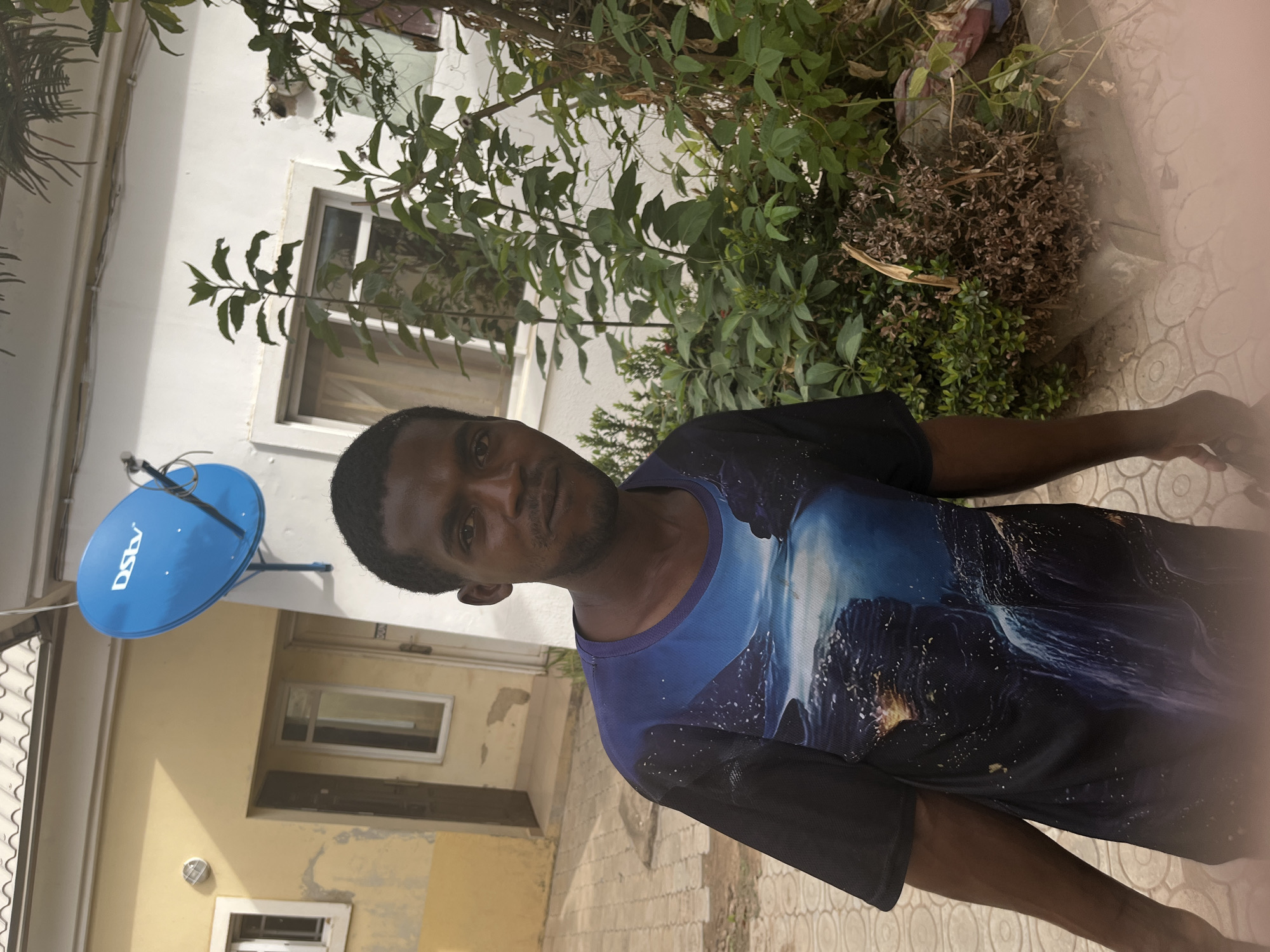 A man stands outdoors in what appears to be a residential area of Abuja, Nigeria. He is wearing a black and blue patterned shirt. Behind him is a yellow-painted building with white window frames. A blue satellite dish with the "DSTV" logo is mounted on the building's exterior. Green foliage and a small brown bush are also visible in the background, partially obscuring a white building with a window. The ground is paved with interlocking pavers. The man is looking at the camera. The image has a slightly washed-out or overexposed quality.