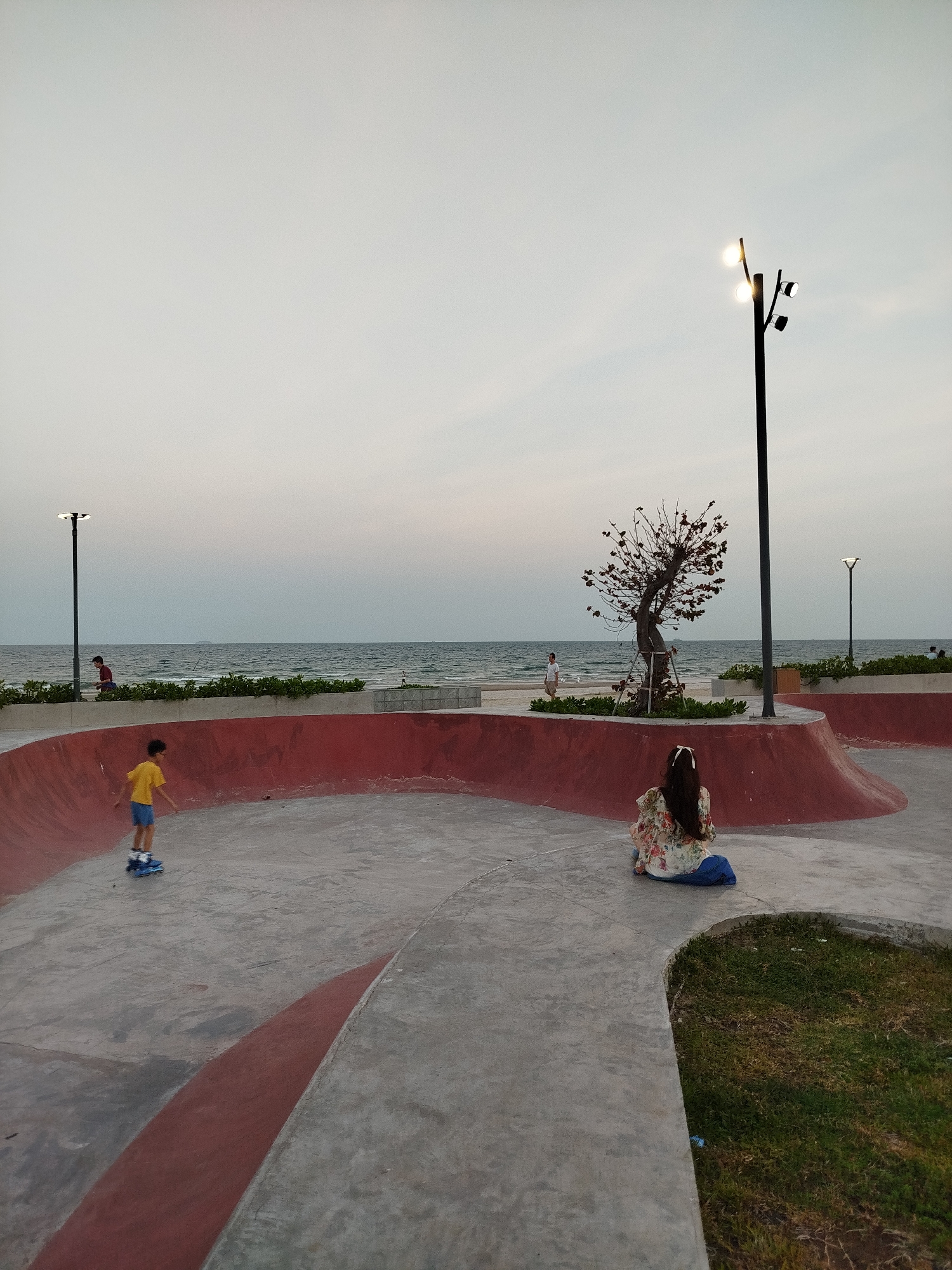 The photograph depicts a skatepark located on the beachfront in Ho Chi Minh City, Vietnam. The skatepark features curved red walls and a grey concrete surface. A young boy is roller skating on the concrete. A woman is sitting on the edge of the skatepark, with her back to the viewer, wearing a floral top and dark pants. In the background, there is a beach with waves and several individuals walking along the shore. Three lamp posts with lights are visible near the beach. A bare tree is situated near the woman. The sky is overcast.