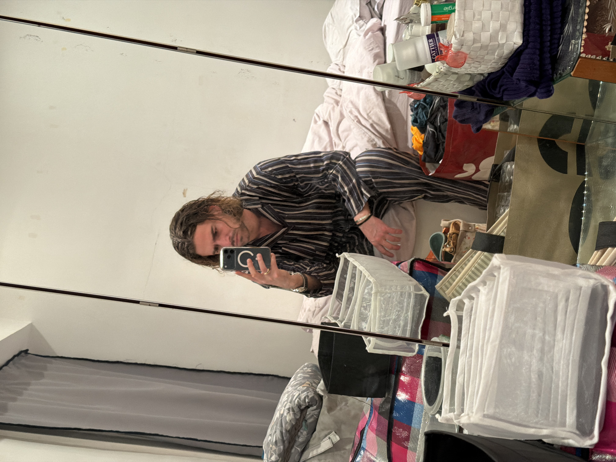 A reflection in a mirror captures a person with long hair wearing striped pajamas, lying on a bed and holding a phone as if taking a selfie. The bed is covered with wrinkled sheets, and an assortment of items like toiletries, bags, and other personal belongings are scattered around the bed and on a shelf beside the mirror. In the lower left corner, curtains are visible, suggesting the interior of a room. There is no indication that this image was taken in Paris, France.
