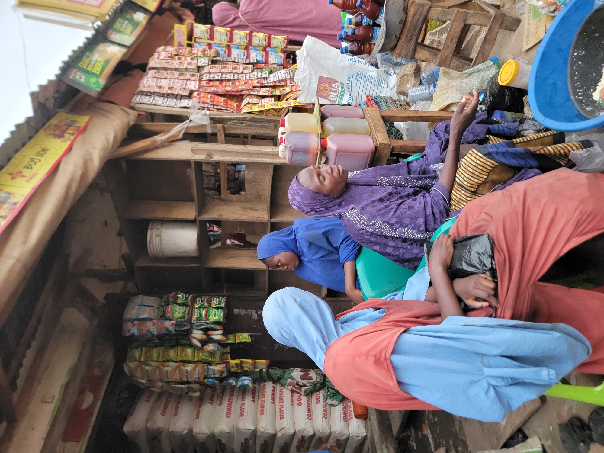 Three women sit amongst goods displayed in a small stall in Jos, Nigeria. One is in a purple hijab and dress, laying back. The second, in a blue hijab, bends forward looking at something on the lower shelf. The third, wearing a blue hijab with a rust-colored dress, is sitting on a green plastic chair, holding a black plastic bag in her hands. The shelves of the shop are stocked with flour, pasta, seasonings, drinks, and other dry goods. The ramshackle shop has a tin roof.