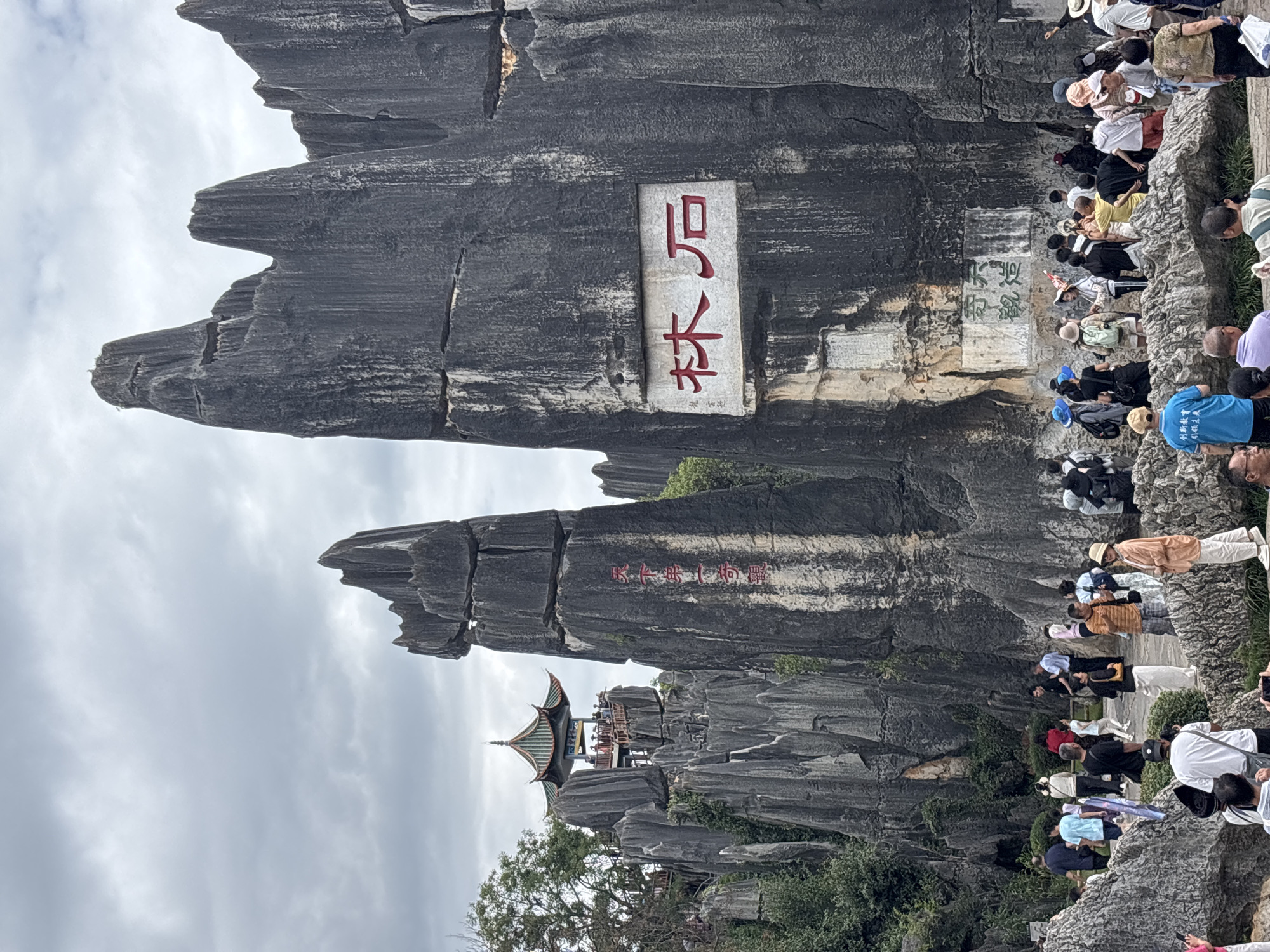 This is a photograph of the Stone Forest National Geopark, located in Kunming, China. The scene depicts a large collection of towering, irregularly shaped limestone formations, commonly referred to as karst formations. In the foreground, a group of tourists are milling around the base of these formations. Some are taking photographs, while others are looking up at the rock structures or interacting with each other.
Dominating the right side of the image is a massive limestone pillar with a rectangular stone plaque inscribed with red Chinese characters, which reads "林石" (Lín Shí), meaning "Stone Forest." To the left, another prominent rock formation rises, also bearing red Chinese calligraphy. Further in the background, perched atop a cluster of rocks, is a traditional Chinese pavilion with a multi-tiered, green-tiled roof. Several individuals are visible on the pavilion's viewing platform, observing the surrounding landscape. The sky is overcast, with a soft, diffused light. The overall impression is of a popular tourist destination showcasing unique geological features.