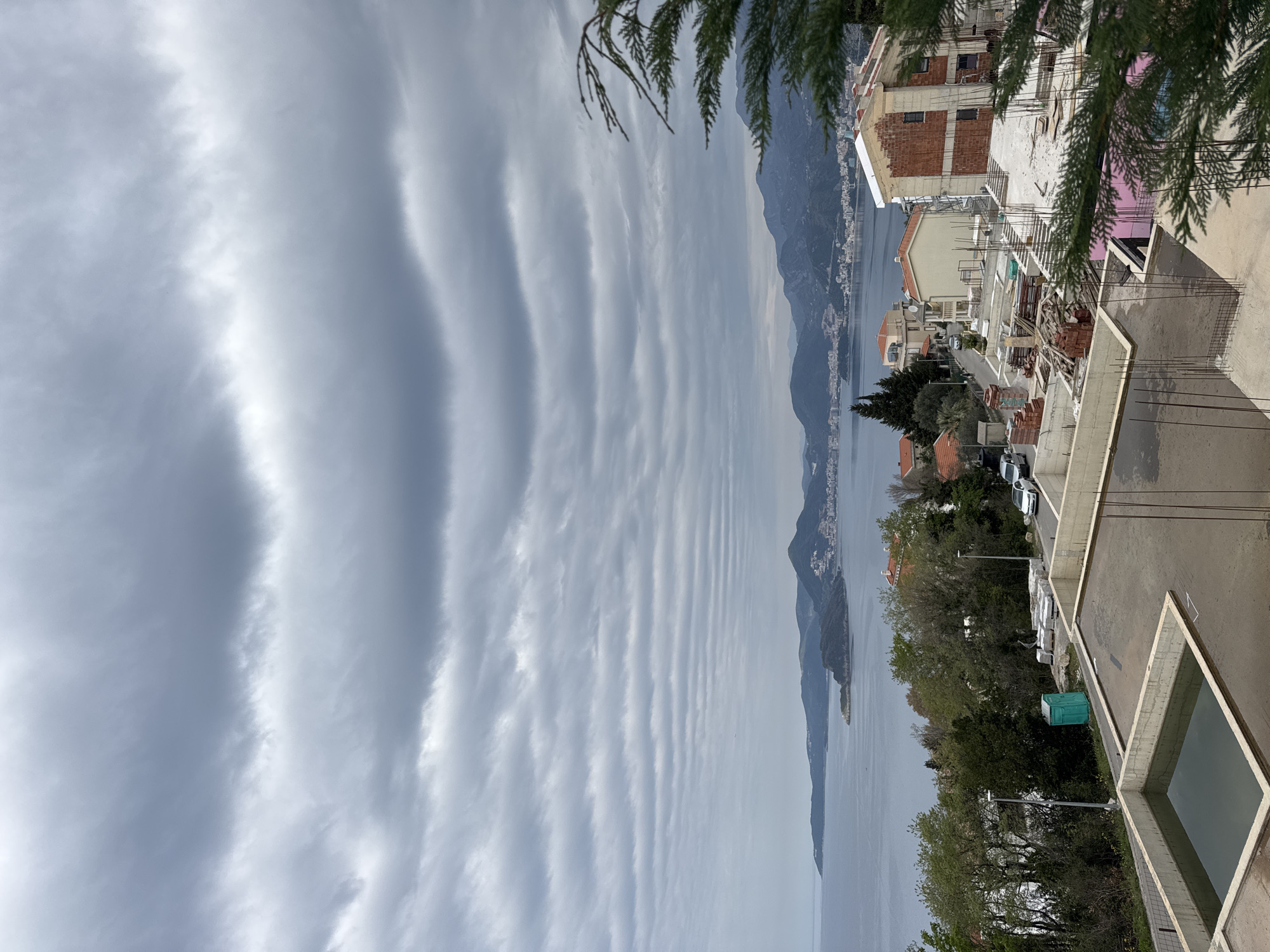 The image captures a view in Blizikuće, Montenegro, featuring a sky dominated by linear, wavy cloud formations stretching across the frame. A body of water, likely the Adriatic Sea, extends towards distant, hazy mountains, with a landmass or island visible in the water. The foreground includes residential buildings and infrastructure under construction, indicative of urban development. Trees are interspersed among the buildings. A portion of a building under construction is visible.