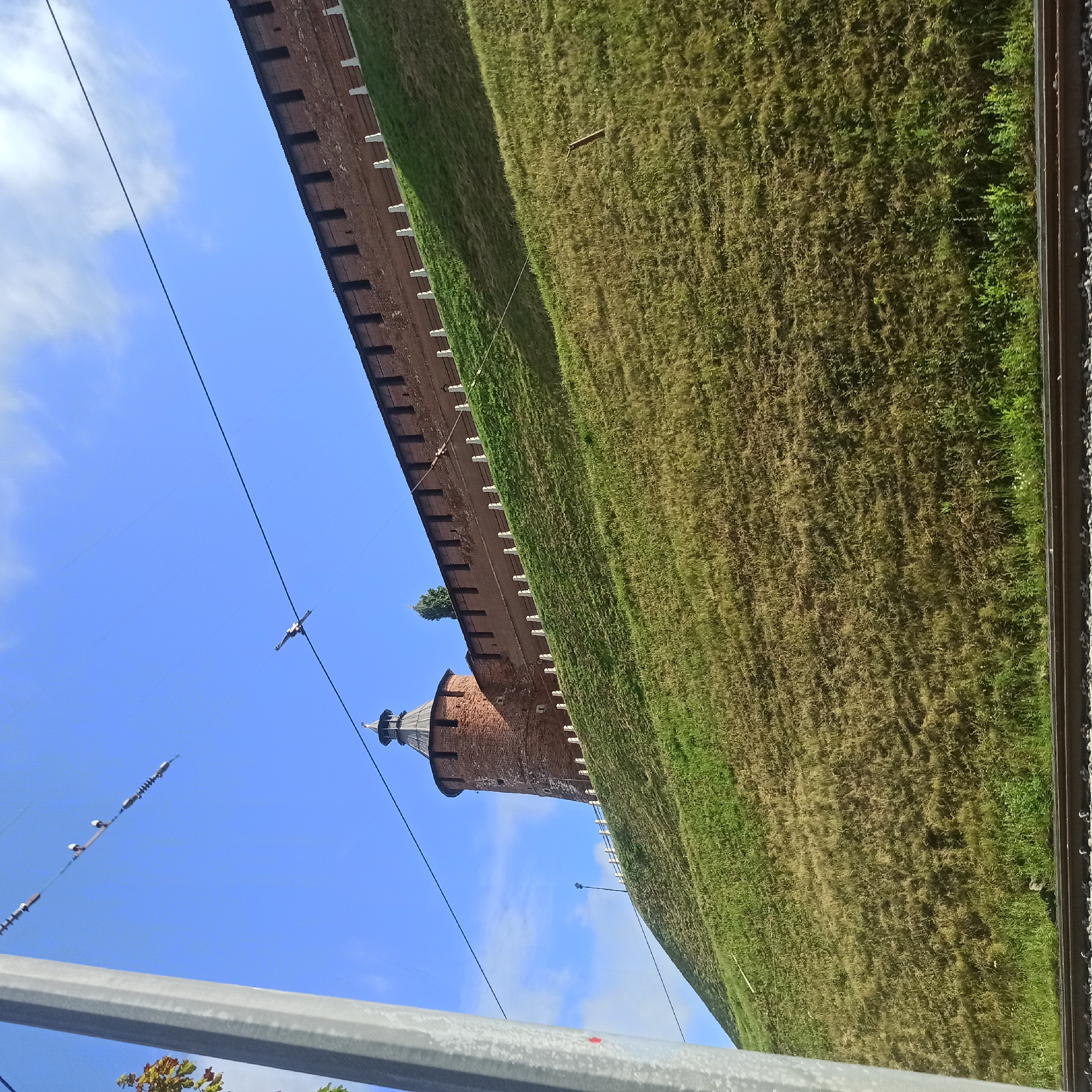 The image depicts a view of the Nizhny Novgorod Kremlin walls and a round tower, also known as a watchtower, in Nizhny Novgorod, Russia. The Kremlin walls, constructed of brick, are visible on a steep, grassy slope under a bright blue sky with scattered white clouds. A white railing with posts runs along the top of the slope, below the Kremlin wall. The round tower, made of reddish-brown brick, is situated on the left side of the image, extending above the slope and partially obscured by the wall. A single tree is visible behind the wall, near the tower. The foreground of the image shows a section of railway tracks, suggesting the viewpoint is from ground level near the tracks. Overhead, power lines stretch across the sky, supported by a tall, grey utility pole on the far left. The lighting suggests it is daytime, with the sun casting shadows.