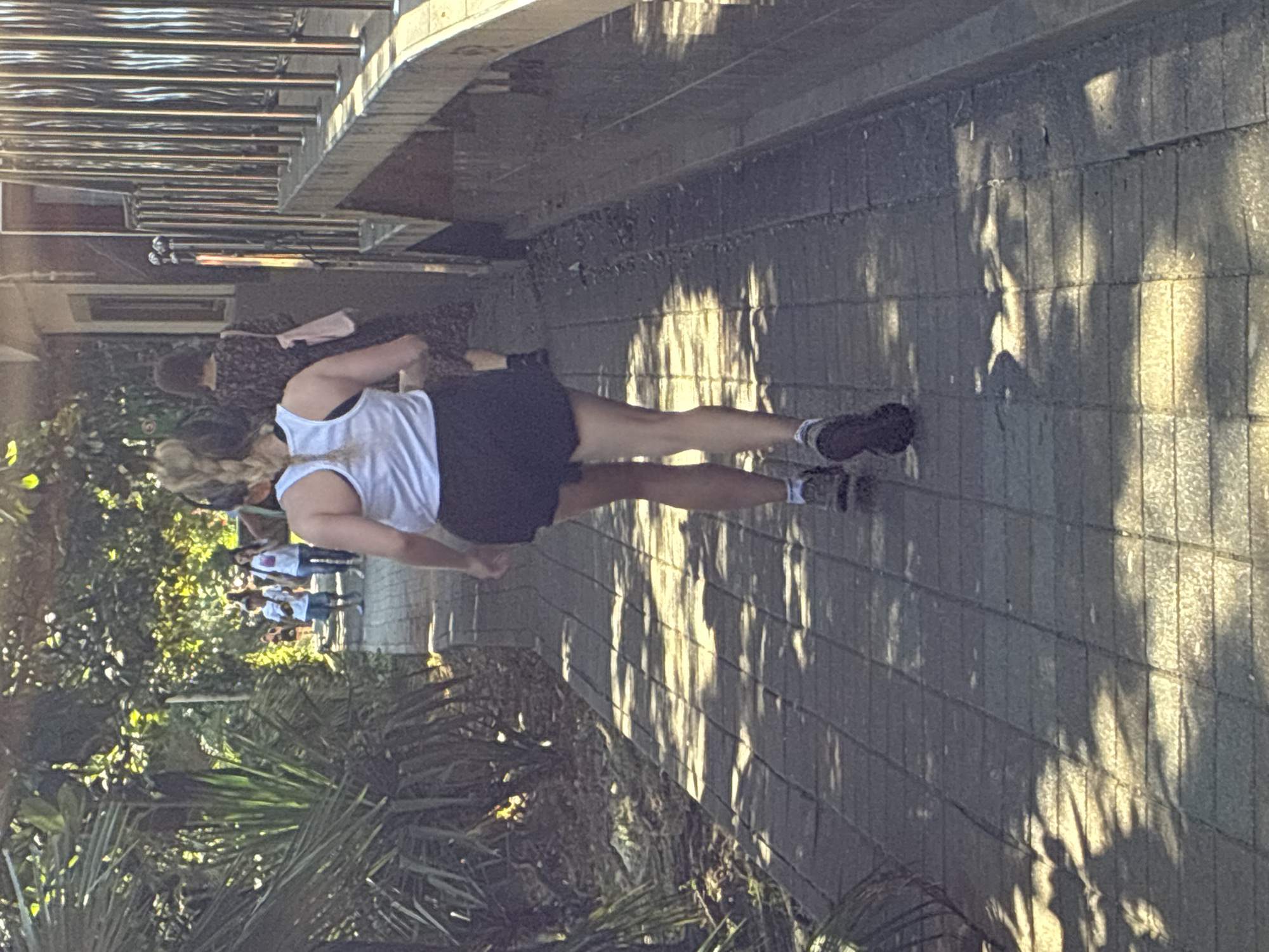 The image captures a sunlit, paved pathway in Sochi, Russia, lined with lush greenery on the left and a building with a metal railing on the right. A woman, viewed from behind, is captured mid-stride as she runs on the pathway. She is wearing a white tank top and black shorts, with her blonde hair styled in a braid. Her athletic shoes are visible. In the background, several other people are walking along the pathway, including a child in a white t-shirt and blue shorts and two women, one carrying a pink bag. The sunlight casts dappled shadows across the pathway, suggesting a warm, pleasant day. The environment appears to be an outdoor recreational area or a promenade in Sochi.