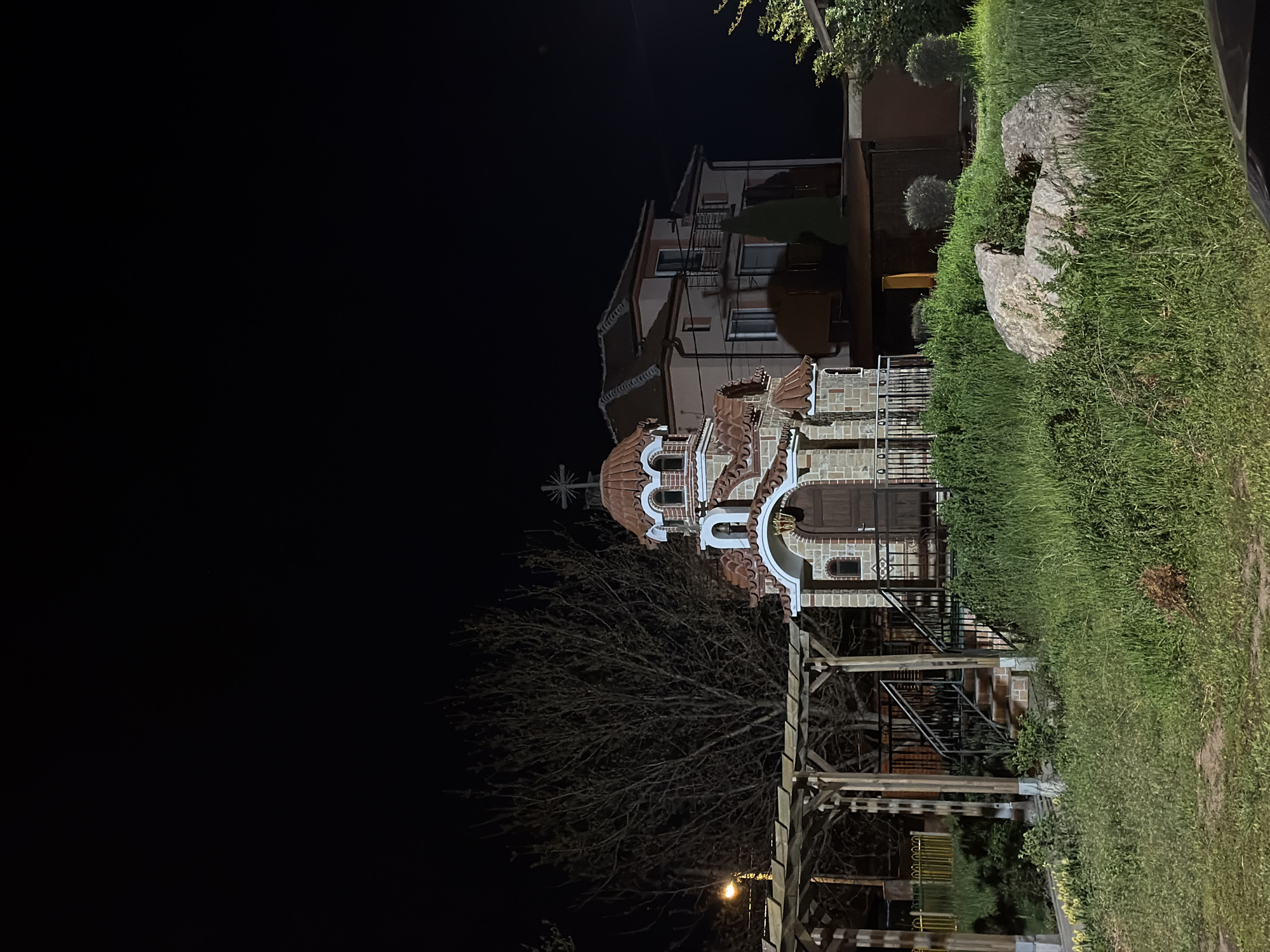 The image shows the Sveti Sedmochislenitsi Church in Sofia, Bulgaria, at night. The church is illuminated and the sky is dark. A bare tree is visible in front of the church, and a wooden pergola-like structure is in the foreground. To the right of the church, a multi-story building can be seen, as well as a grassy area with rocks. The image is taken from a low angle.