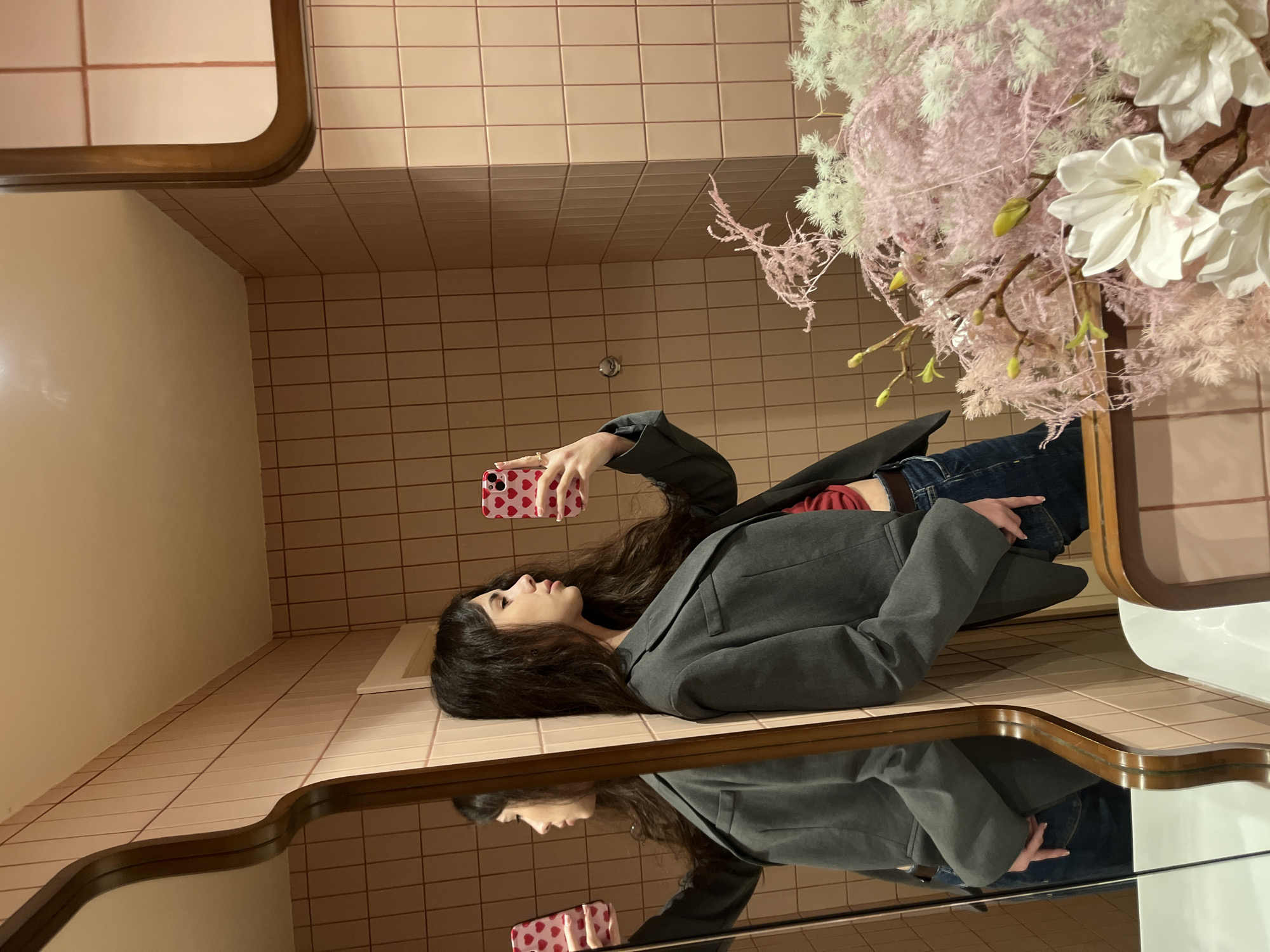 A young woman with long dark hair, wearing a grey blazer over a red top and dark jeans, leans against a pink tiled wall while taking a mirror selfie with a phone in her right hand. The phone case is pink with red hearts. She is positioned in the left half of the frame, facing the mirror. To the right of the frame, a large floral arrangement with pink and white feathery branches and white flowers is visible. The background is composed of pale pink square tiles. The lighting is warm and ambient.