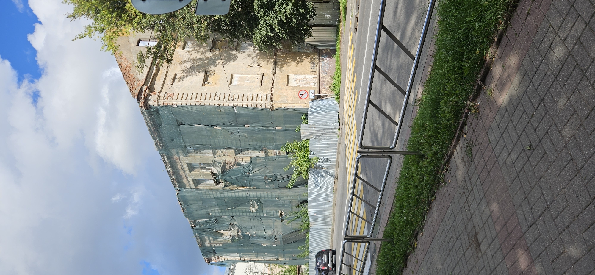 The image depicts a building undergoing renovation or demolition in Vitsebsk, Belarus. The facade of the building is covered with a large green tarp, obscuring much of its architectural details. The building appears to be old, with visible brickwork and signs of decay. In front of the building, there is a metal fence and some greenery. A road with a pedestrian crossing is visible in the foreground, and a car is driving on the road. A metal railing separates the sidewalk from the road. The sky is partly cloudy with patches of blue.