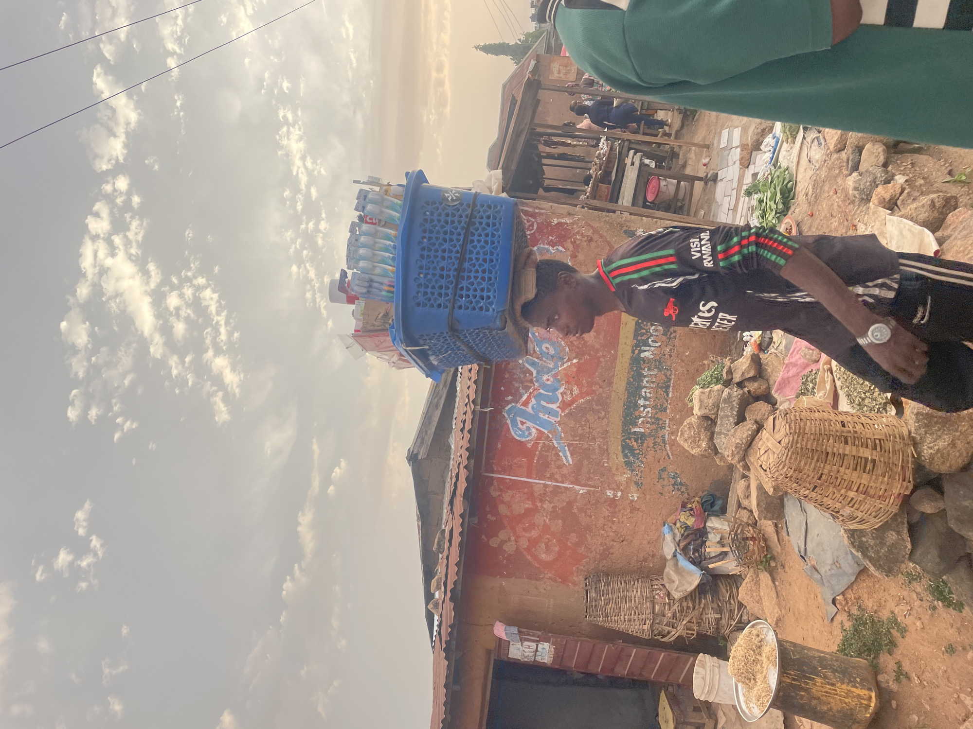 In a busy outdoor marketplace in Jos, Nigeria, a slender man is seen in profile carrying a blue plastic basket laden with toothbrushes on his head. He wears a black jersey with green and red stripes on the sleeves, black track pants, and a watch on his left wrist.
Behind him, the weathered wall of a building displays faded advertisement text. A wicker basket sits on the ground amidst rocks and debris. Other vendors and shoppers are visible in the background. The sky is overcast.