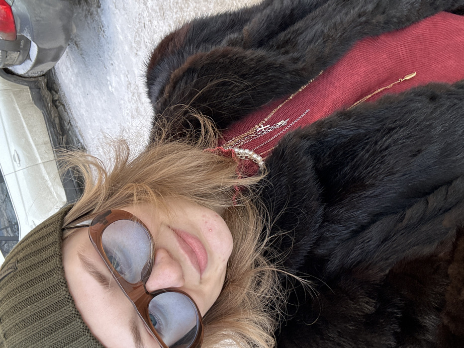 An individual is depicted in a close-up, outdoor, upward-angled selfie taken in Kazan, Russia. The subject, appearing female, has light brown hair, wears an olive green ribbed knit beanie, and large brown-framed sunglasses with reflective blue lenses. A slight smile is present. She is dressed in a dark brown fur coat, under which a red turtleneck garment is visible. Layered jewelry adorns her neck, including a pearl choker and several metallic chain necklaces; two chains feature cross pendants, and one has a small, golden, feather-shaped pendant. The background reveals a snow-covered or icy ground surface, with two vehicles partially visible. A light-colored SUV or sedan, covered in dirt and slush, is parked on the left, and a darker vehicle, also snow-dusted, is partially visible to its right.