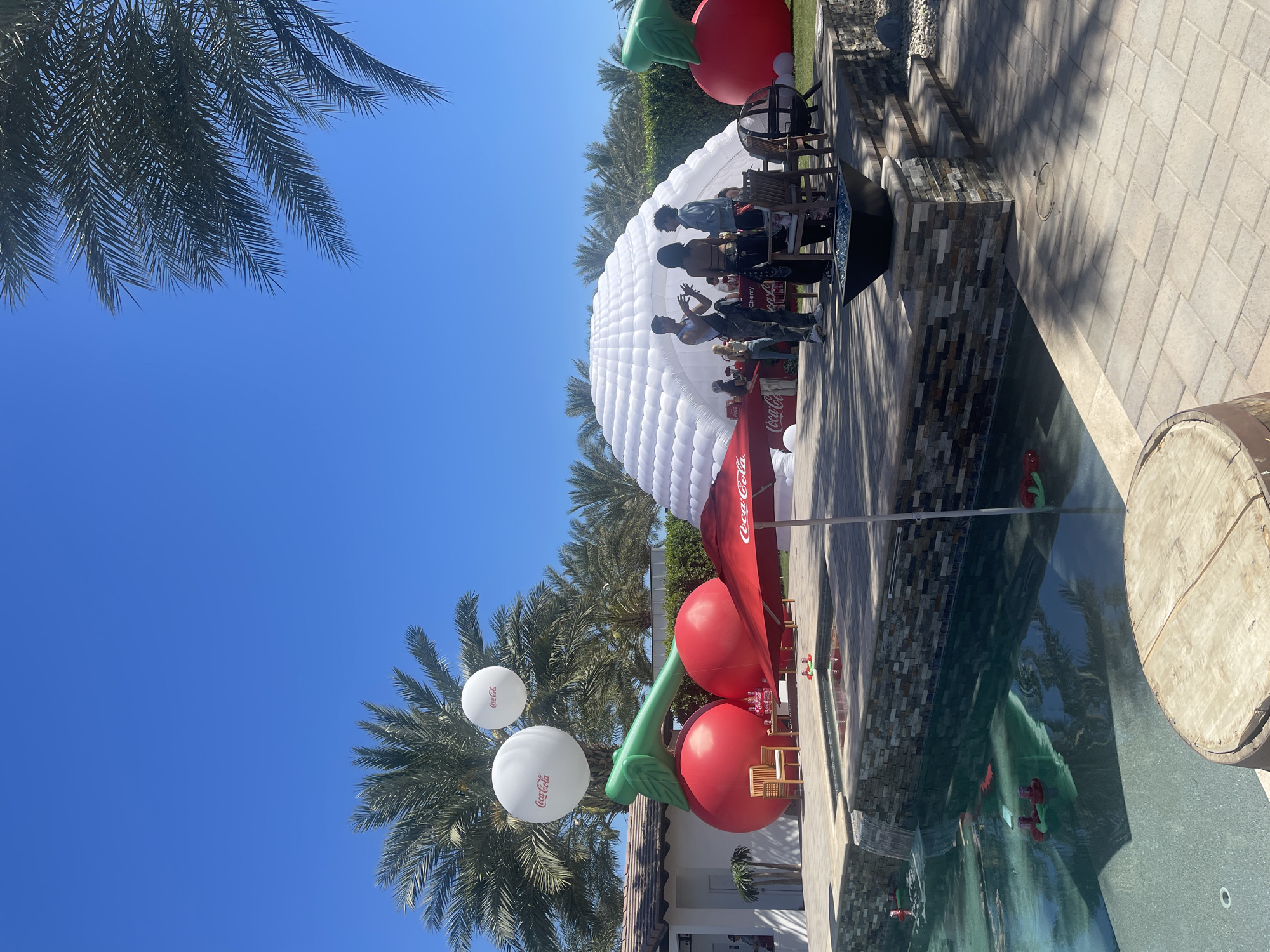 Here's a detailed description of the scene:
**Scene Overview:** The image captures an outdoor event or gathering, possibly a promotional event, on a bright sunny day in Thermal, United States.
**Environment:** Palm trees line the area, indicating a warm climate. There is a pool present, with some possible decorations within it. The ground appears to be a combination of stone pavers and grassy areas.
**Objects:**
* **Large Inflatables:** Several large inflatable structures dominate the scene. There is a large white dome-shaped inflatable, which might be a tent or display area. There are also two large inflatable cherries, a Coca-Cola umbrella, and Coca-Cola balloons.
* **Furniture:** There are chairs near the white dome and Coca-Cola umbrella.
* **Branding:** Coca-Cola branding is prominent throughout the scene, displayed on balloons and an umbrella.
* **Wine Barrel:** A wooden wine barrel is present near the pool.
**People:**
* There are a small number of people gathered near the white inflatable dome. They appear to be interacting with each other.