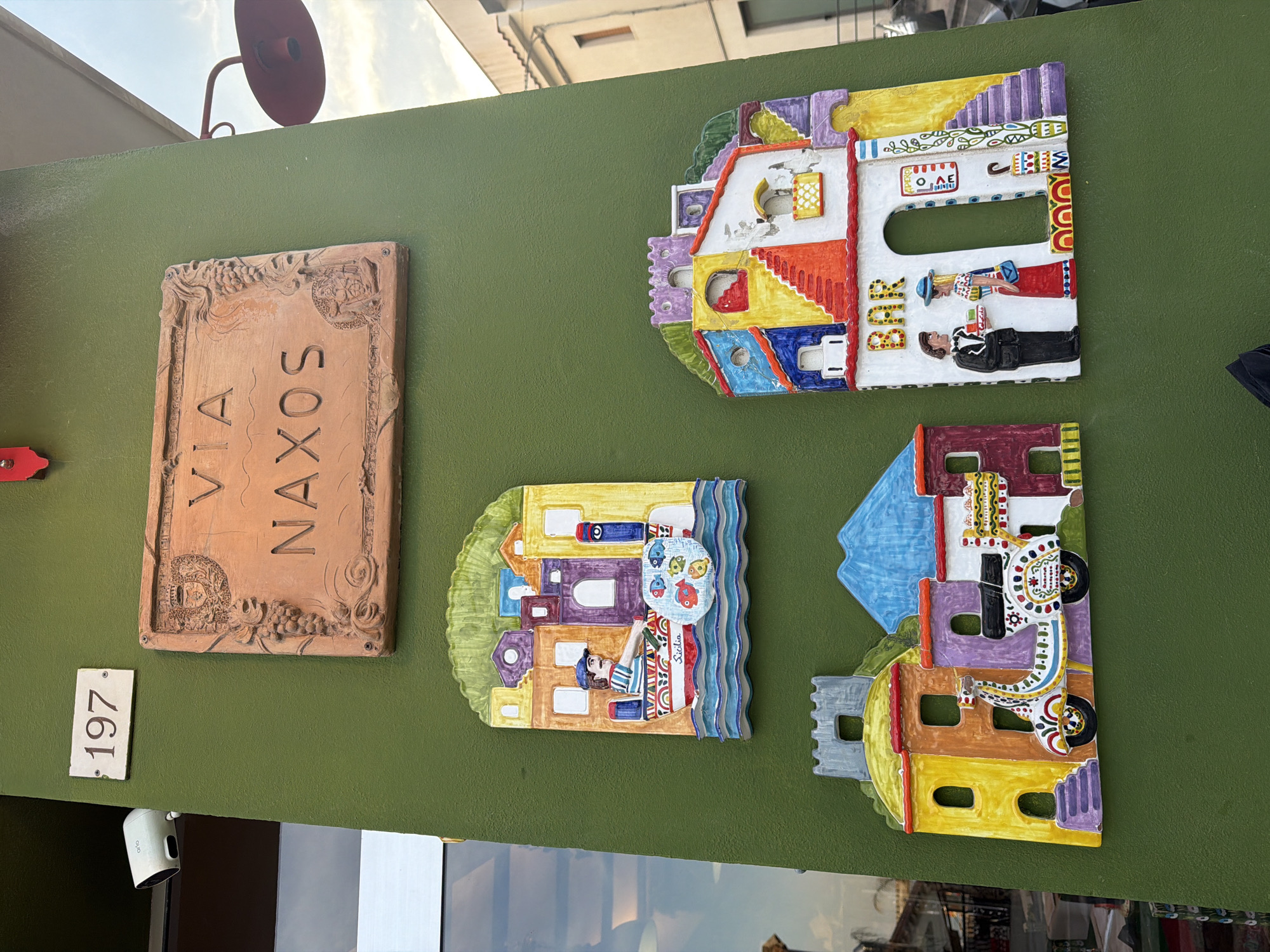 On a green wall, three colorful, stylized ceramic wall hangings and a terracotta plaque are displayed. To the left, a rectangular plaque reads "VIA NAXOS" with ornate floral and heraldic designs. Adjacent, a small white plaque shows "197". The top-right ceramic piece depicts a building with figures, one in a suit and the other in a dress. Below, another depicts a person selling fish, and a third piece depicts a decorated vehicle. The scene appears to be from the city of Chianchitta, Italy. A security camera is visible below the "197" sign. The red light fixture above the ceramic wall hangings is also included in the image.