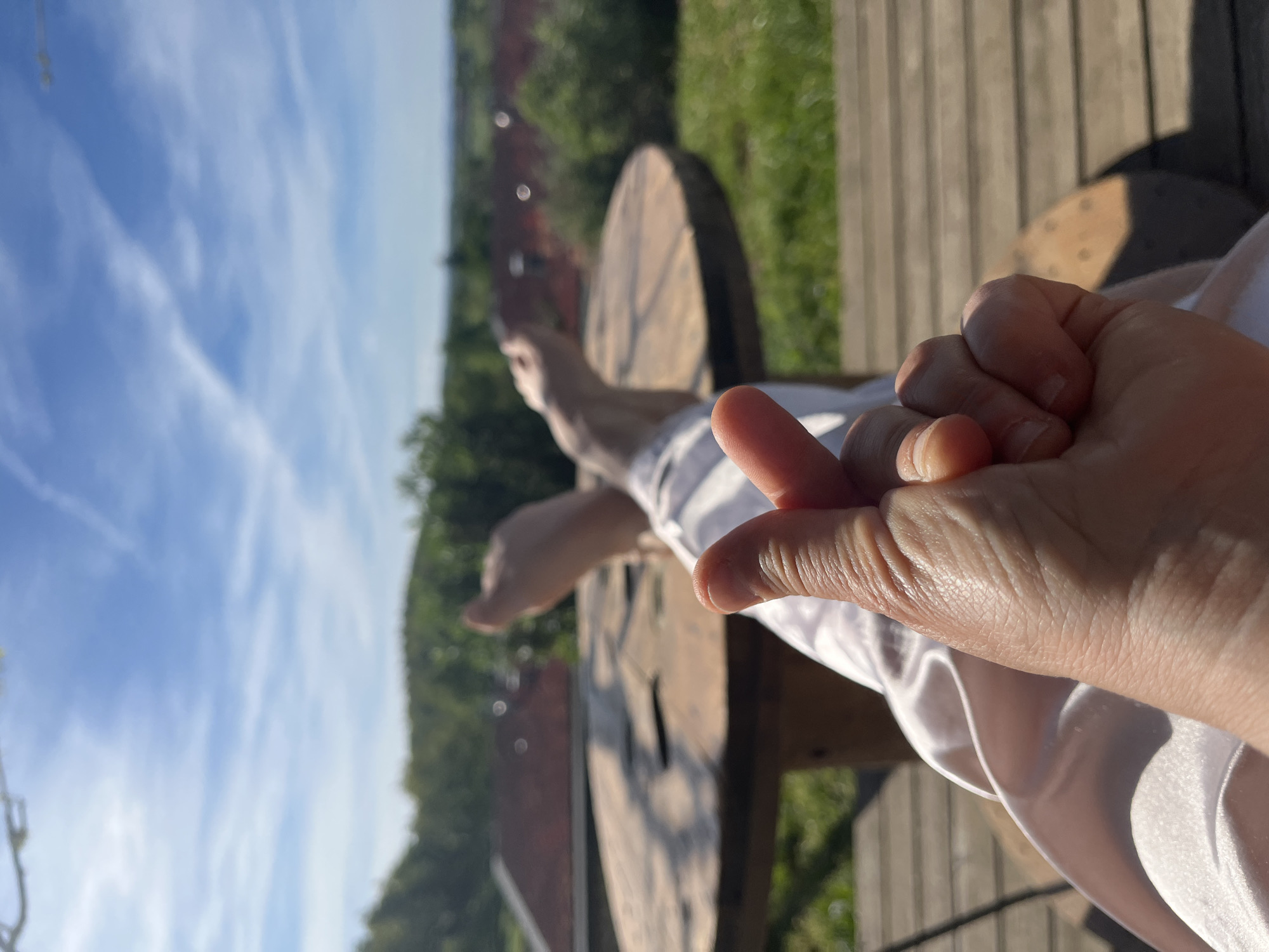 The image depicts a bright outdoor scene, likely in Chambœuf, France, under a clear blue sky with scattered white clouds. In the foreground, a person's hand is visible on the right, making a "finger heart" gesture. The hand is well-lit by bright sunlight, highlighting skin textures and creases. The person is wearing a white garment, possibly made of satin, which is visible around the wrist and lower arm.
Further into the midground, the person's bare feet can be seen in a relaxed position, resting on or near a large wooden spool, which appears to be repurposed as outdoor furniture like a table or seat. The background consists of lush green foliage, suggesting trees and grass, and a wooden fence or structure. The sun casts distinct shadows, indicating a sunny day, likely morning or afternoon. There is no visible text in the image. The overall impression is one of relaxation and affection in a natural, serene environment.