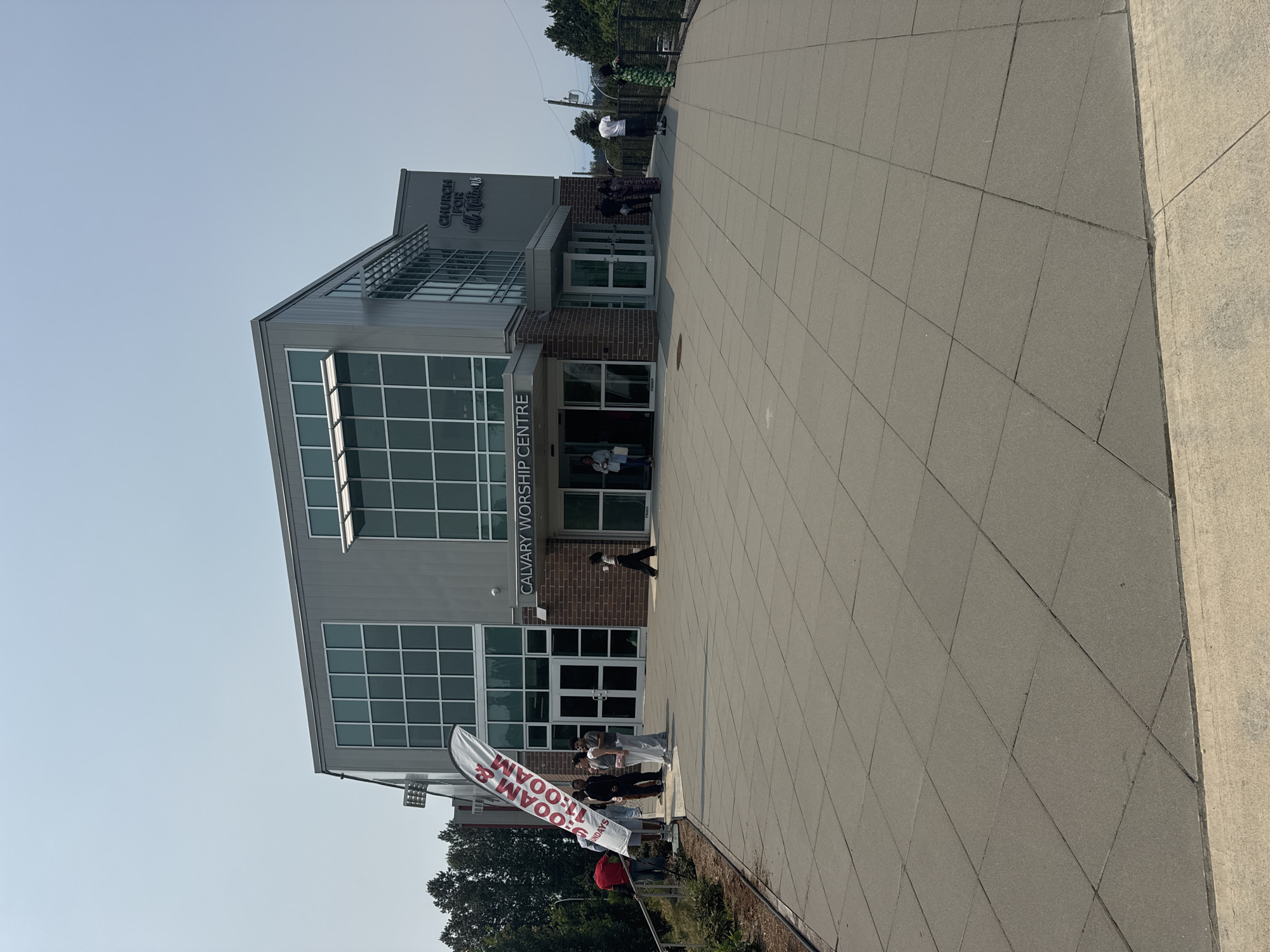 The image shows the Calvary Worship Centre, a church located in Surrey, Canada. The building is modern with a grey facade and large windows. In front of the entrance, there is a paved walkway with several people gathered. A tall white banner with red lettering is visible on the left side of the entrance, advertising "9:00 AM & 11:00 AM Sundays". The sky is clear and the weather appears to be warm.