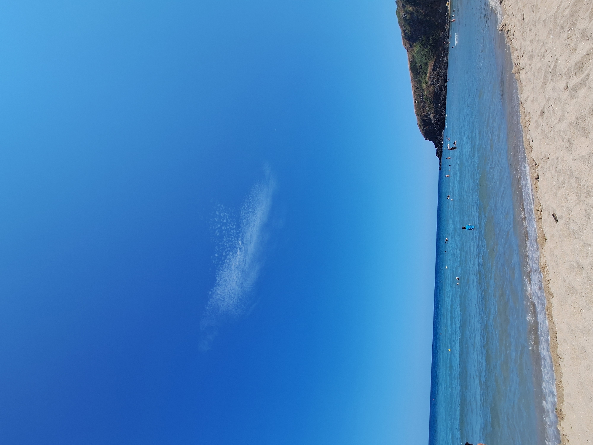 The image depicts a sunny day at a beach in Sinemorets, Bulgaria, likely during the summer. A clear, vibrant blue sky dominates the upper half of the frame, with a small, wispy cloud formation visible in the upper left quadrant. The lower half of the image shows a tranquil sea with gentle waves lapping onto a sandy beach. The water is a beautiful gradient of blues, from light turquoise near the shore to a deeper blue further out.
Several people are seen enjoying the water, swimming and wading. Some are further out, while others are closer to the shore. A rocky, verdant headland juts out into the sea on the right side of the image, providing a natural backdrop. The sand in the foreground appears light and fine-grained. The overall atmosphere is one of peaceful recreation and natural beauty.
