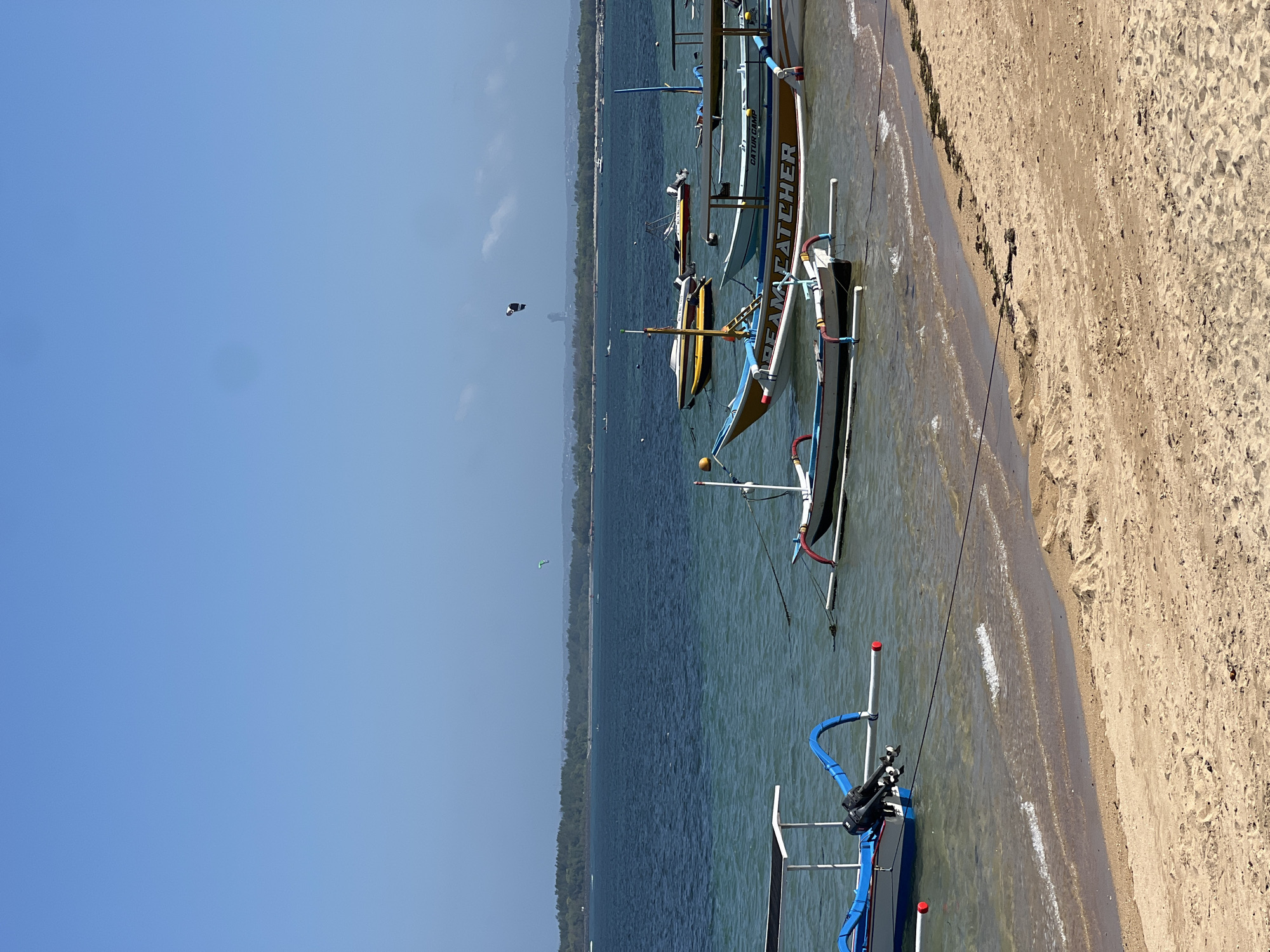 This is a daytime, eye-level, medium shot of a beach in Denpasar, Indonesia, likely Sanur Beach. The foreground features a sandy beach with some debris. The midground is occupied by the calm, shallow, turquoise ocean with several traditional Indonesian outrigger boats (Jukung) moored or anchored. The boats are of various sizes and colors, some with outboard motors. One prominent boat in the center has the word "DREAM CATCHER" written on its side in yellow lettering. In the background, a distant, tree-lined shore is visible under a hazy, light blue sky. Two figures are seen kiteboarding in the distance, identifiable by their colorful kites. The water is clear enough to see the sandy bottom in some areas near the shore.