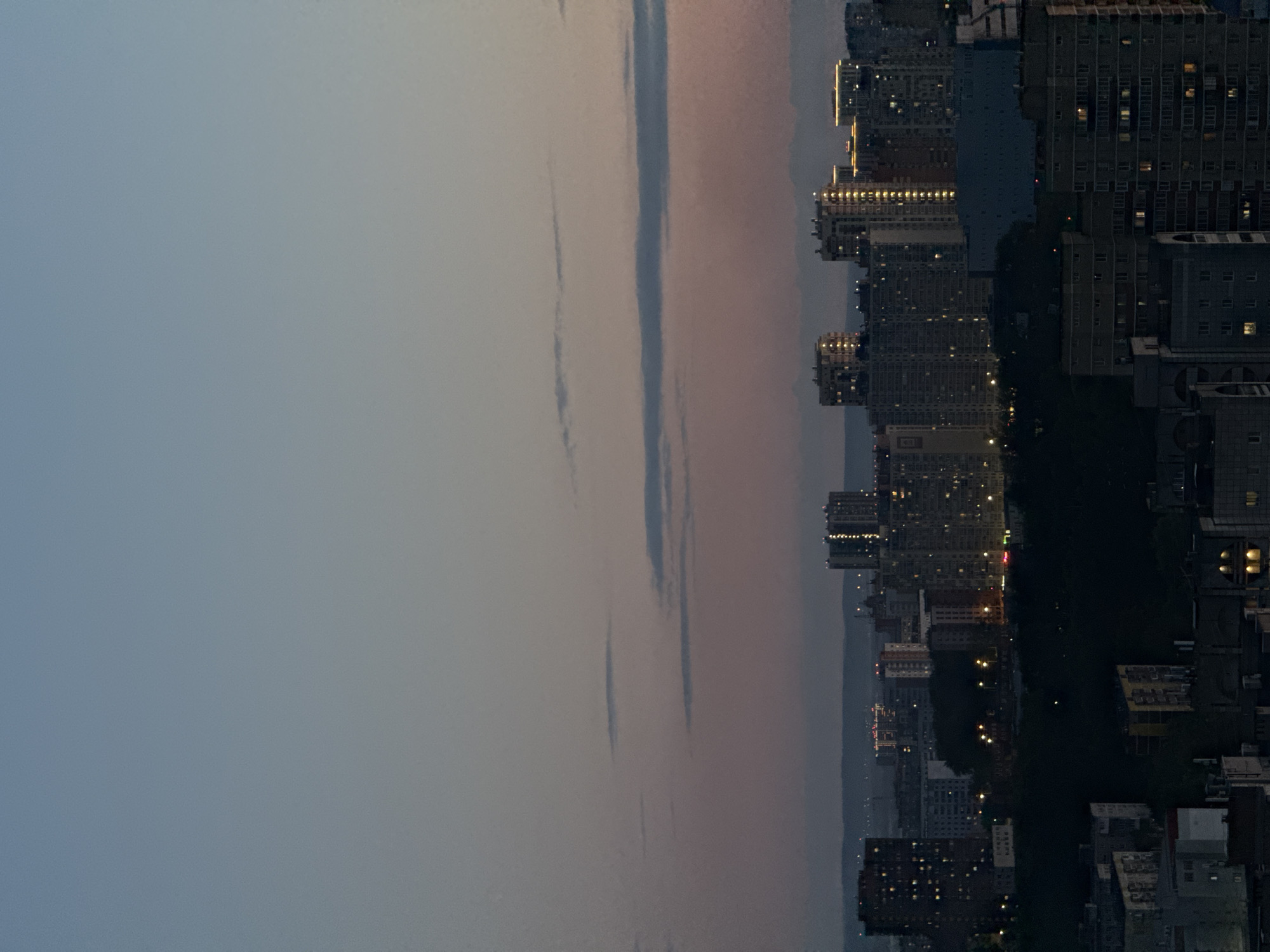 An elevated view captures a city skyline at dusk or dawn. The upper two-thirds of the image depict the sky, transitioning from a soft blue-grey at the top to a warmer, subtle pinkish-orange closer to the horizon. Several elongated, wispy clouds stretch horizontally across the sky.
The lower third of the frame is filled with numerous urban buildings, predominantly high-rise structures. Many windows in these buildings are lit with warm, yellow lights, signifying evening or early morning. A broad, dark expanse, likely a river or a large park, horizontally bisects the urban landscape, separating the clusters of buildings. Beyond this dark feature, distant hills or landforms are faintly visible on the horizon.
No individuals or specific events are discernible. The image conveys the peaceful transition of a city between day and night, characterized by the interplay of natural light and artificial illumination. The weather appears clear with minimal cloud cover. The scene represents a view of Perm, Russia. No text is visible in the media file.