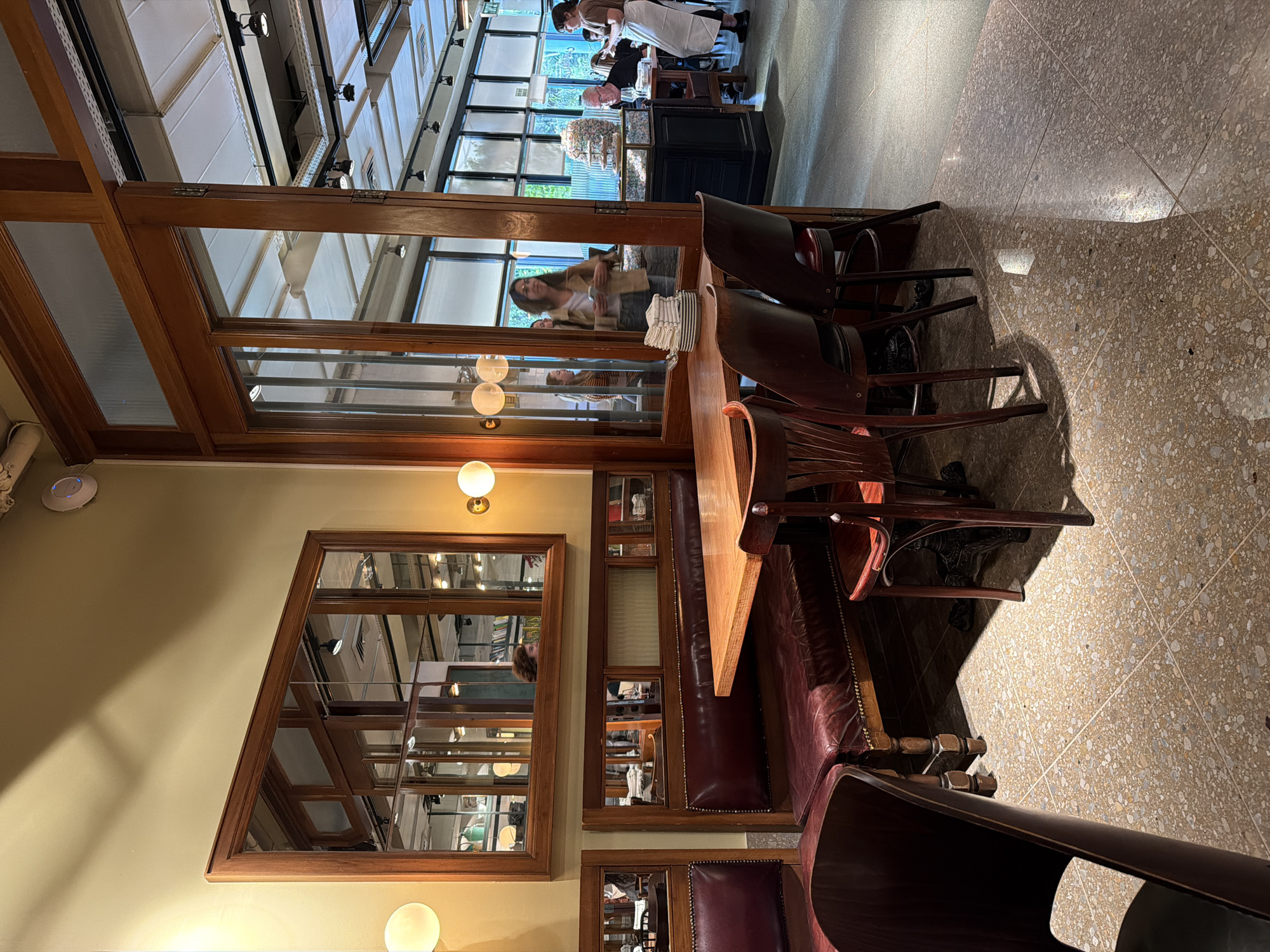 The image shows the interior of a restaurant or cafe in Tel-Aviv, Israel. The space features a combination of modern and vintage design elements. Patrons are seated at tables, and a server is visible in the background. Architectural details include wooden window frames, mirrors, and a high ceiling with a glass roof, suggesting a renovated historical building. The color palette is warm, with light-colored walls, dark wood furniture, and reddish-brown leather seating. The floor is a textured, speckled pattern. The ambiance appears relaxed and upscale.
