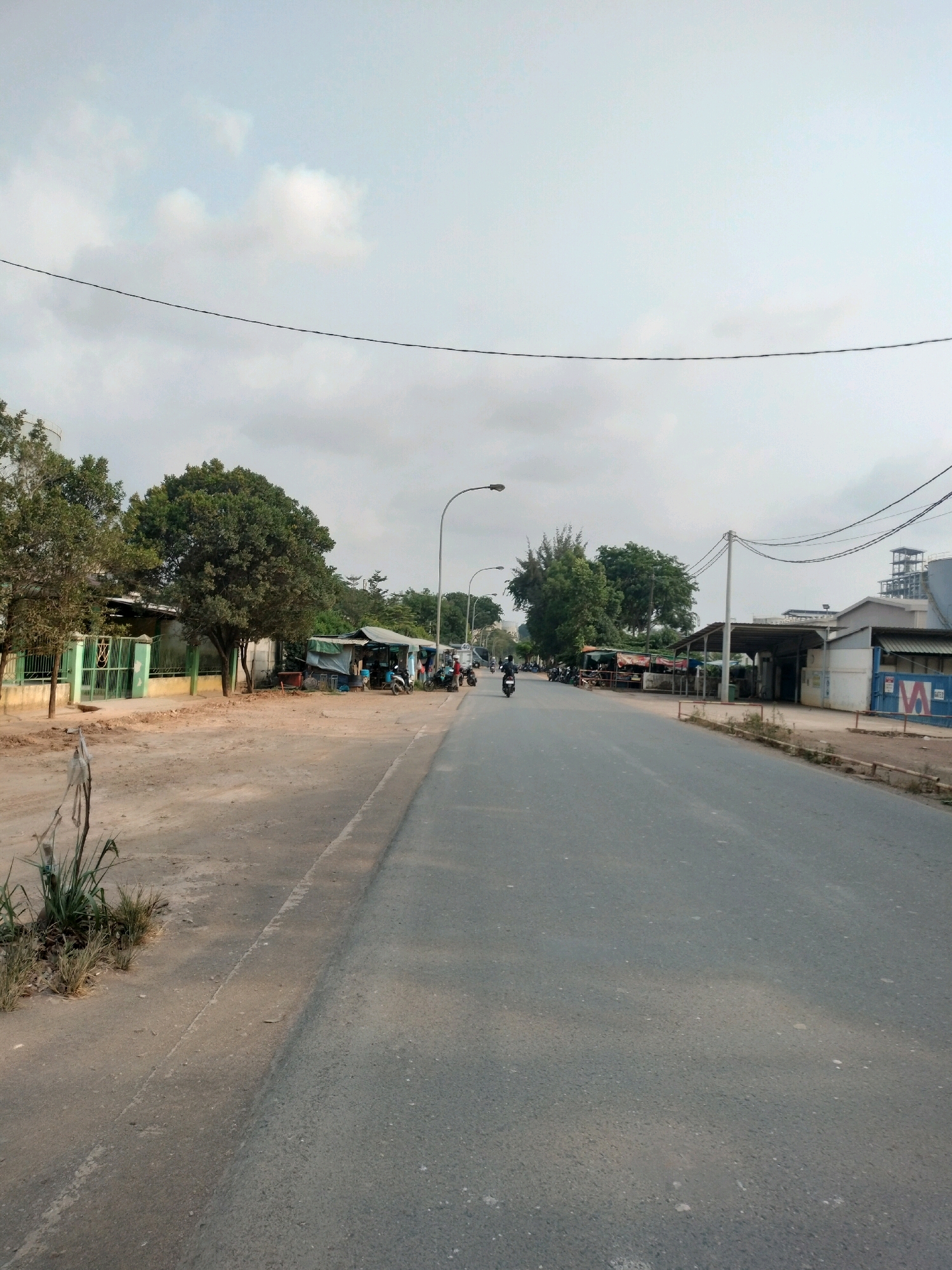 This is a photograph of a street in Batam, Indonesia, likely in an industrial or commercial area. The road is paved and appears to be a two-lane street. On the left side of the road, there is a dirt shoulder with some vegetation and what appears to be a green fence with a gate leading to a building. Further down the left side, there are several small, makeshift structures with awnings, and a few motorcycles are parked. A person is visible near one of these structures. On the right side of the road, there are industrial buildings, including a larger structure with multiple levels in the background. Overhead, there are power lines and a street light. The sky is overcast with scattered clouds. A few motorcycles are seen on the road, one with a rider. The overall impression is a functional, somewhat unkempt, commercial or industrial zone.