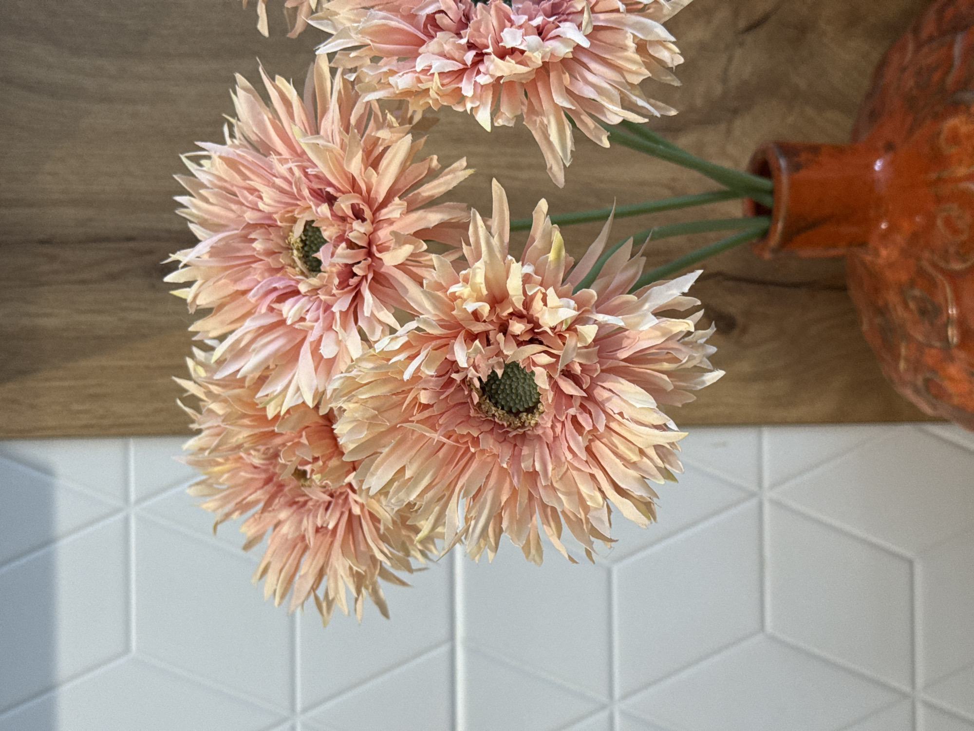 A close-up, indoor vertical shot displays a bouquet of artificial flowers in an orange ceramic vase. The flowers, resembling gerbera daisies, feature numerous thin petals transitioning from pale peach to light pink at their centers, which are dark green. There are at least five visible flower heads, arranged with green stems extending into the vase. The vase itself is textured with an embossed pattern and has a rustic, reddish-orange glaze. Behind the vase and flowers, the background is divided vertically. To the left, a white wall exhibits a three-dimensional geometric pattern composed of interlocking diamonds or hexagons. To the right, a wooden surface with a distinct vertical grain provides a contrasting backdrop. The lighting appears soft, illuminating the petals and highlighting the textures of the wall and vase. No people are present.