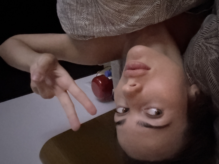 A woman is pictured upside down, making a peace sign with her left hand. She is partially covered by a patterned blanket, which frames her face. Her gaze is directed to the right. A red object sits in the background, near her shoulder, while a white and brown surface appears below her. The image is dimly lit, and the background is mostly black.