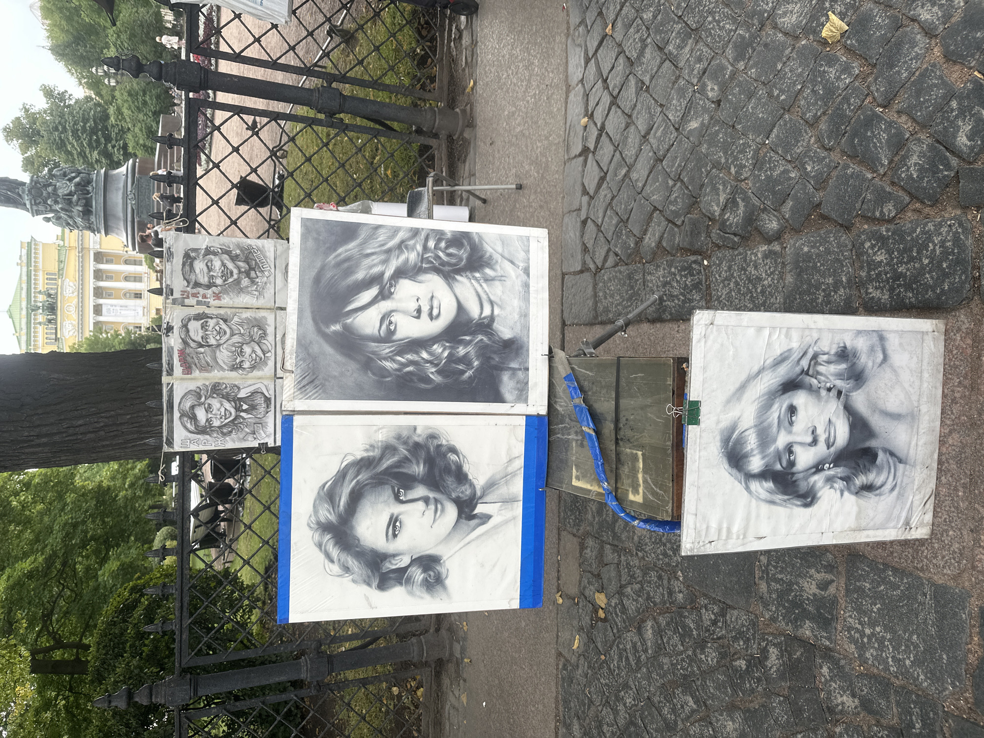 An outdoor display of charcoal portraits is set up on a cobblestone sidewalk in Saint Petersburg, Russia. In the foreground, three large framed portraits of women are displayed. The leftmost portrait shows a woman with voluminous curly hair and bright eyes. The middle portrait is of a woman with wavy dark hair and a serious expression. The rightmost portrait depicts a woman with blonde hair and pearl earrings. Above these, four smaller caricatured portraits of men and women are displayed on a stand. Behind the portraits, a black metal fence separates the sidewalk from a park area with trees and benches. In the background, a large, ornate yellow building with columns and a dome is visible, along with a monument. The scene suggests a public space where artists display their work for sale to tourists or locals.