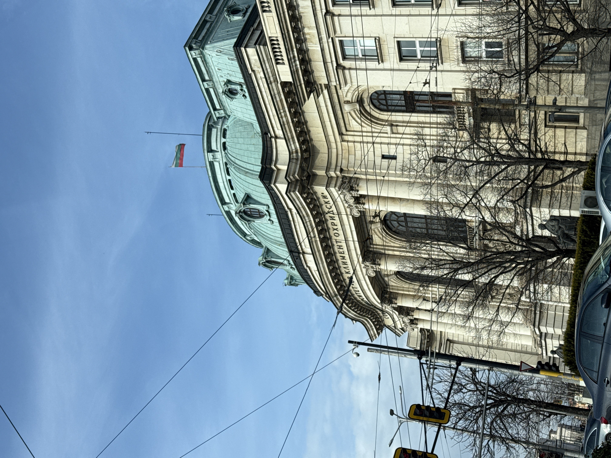 The image captures the National and University Library "St. Kliment Ohridski" in Sofia, Bulgaria, taken from a low angle. The library's architectural features are prominent, including its dome, windows, and the inscription "Св. Климент Охридски" in Cyrillic. A Bulgarian flag flies atop the building. Power lines crisscross the sky, and bare tree branches are visible against the building's facade. In the foreground, traffic lights and a portion of a parked car suggest an urban setting. The sky is clear and blue, adding to the brightness of the scene.