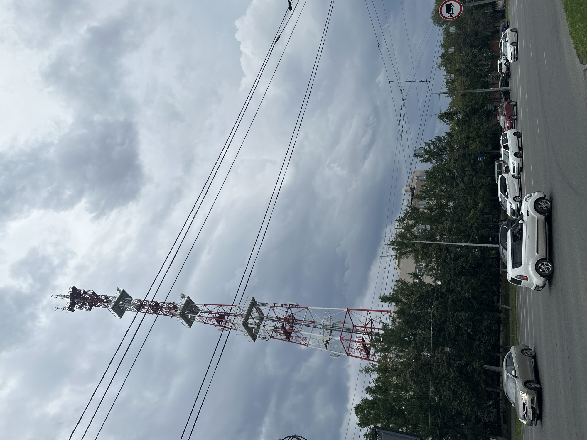 A tall, red and white telecommunications tower stands prominently against a cloudy, overcast sky. Several power lines run diagonally across the frame, intersecting the view of the tower. The tower is situated behind a line of lush green trees, beyond which is a low-rise building. In the foreground, a multi-lane asphalt road is visible with several vehicles in motion. A silver sedan is in the left lane, while a white compact SUV is in the right lane. Other cars are also present further down the road. A traffic sign depicting a truck within a red circle and slash is visible on the right side of the frame.