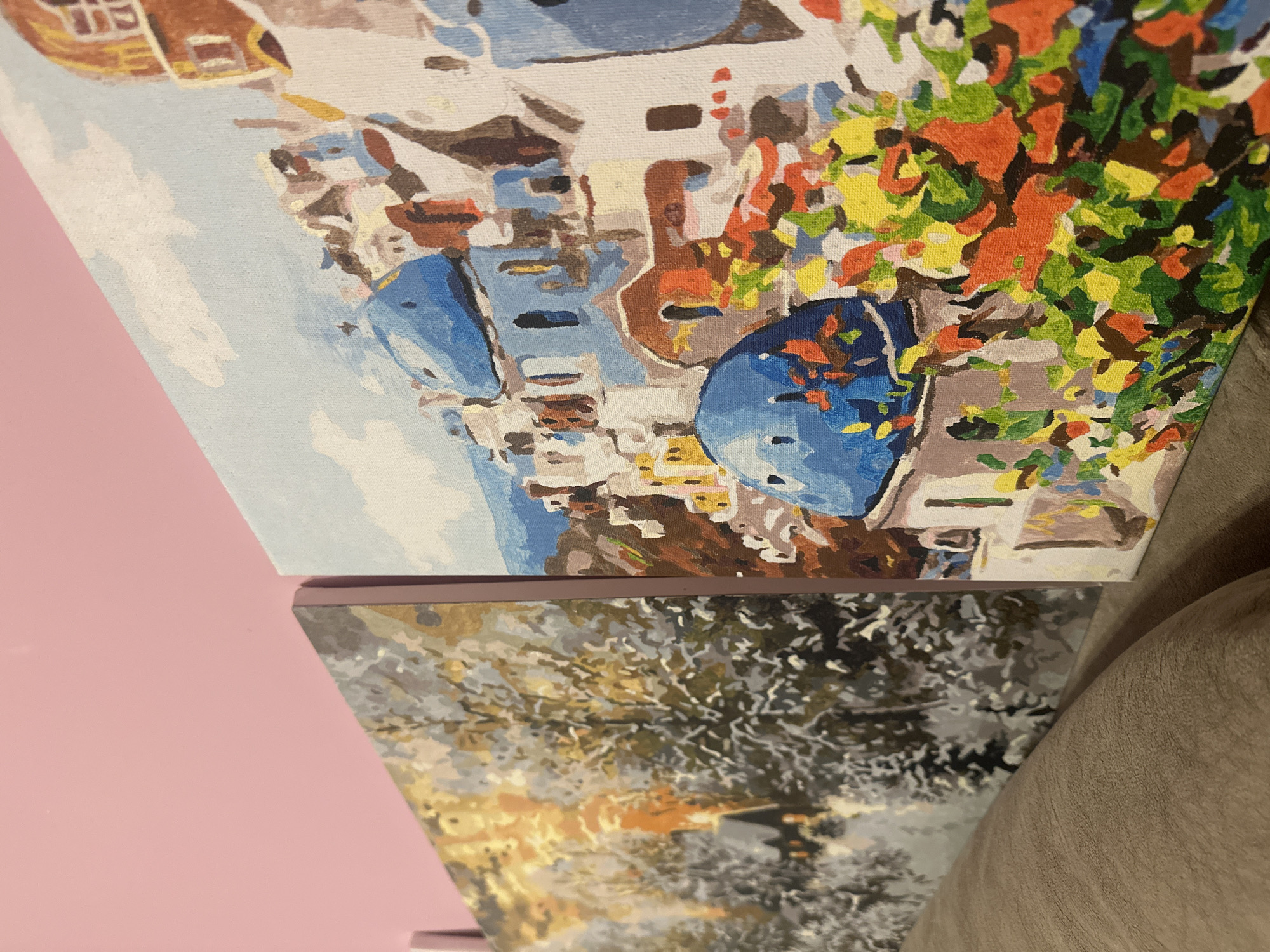 Two paintings are stacked. The top painting depicts a coastal scene with colorful flowers, white buildings, and blue accents, reminiscent of Santorini, Greece. The lower painting shows a winter scene with bare trees and a blurred background. The background consists of a pink wall and a beige couch. The location is likely a home interior, but the depicted scenes in the paintings are not in Varna, Bulgaria.