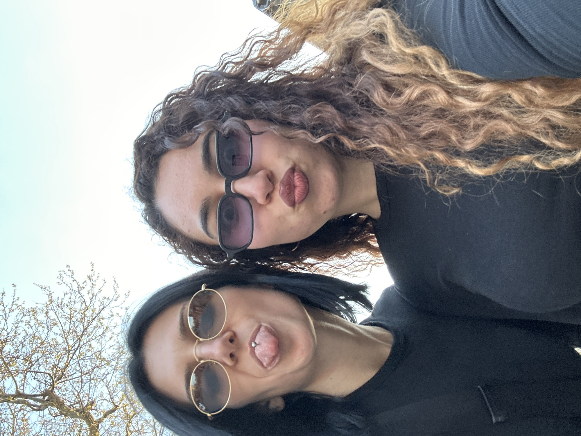 Two women are captured in a playful, low-angle selfie, looking down towards the camera. The image is oriented with the top of their heads towards the right side of the frame and their bodies extending towards the left, set against a bright, clear sky.
The woman in the foreground, positioned slightly to the bottom and left, has dark, straight hair that frames her face. She wears round, gold-rimmed sunglasses with dark lenses. Her expression is humorous, as she sticks her tongue out, revealing a silver barbell piercing through her tongue. She is wearing a dark, possibly black, casual top or t-shirt.
Behind and slightly above her, the second woman has voluminous, curly brown hair with lighter, golden-blonde highlights. She wears rectangular, dark-framed sunglasses with tinted lenses. Her lips are pursed in a slight pout, adding to the playful mood. She is also dressed in a dark, possibly black, top or shirt.
The background is predominantly a bright, light blue sky, suggesting an outdoor setting on a clear day. To the far left, bare tree branches with emerging light green buds are visible, indicating early spring. No text is visible in the image. The overall mood is lighthearted and friendly, possibly taken in Burgas, Bulgaria.