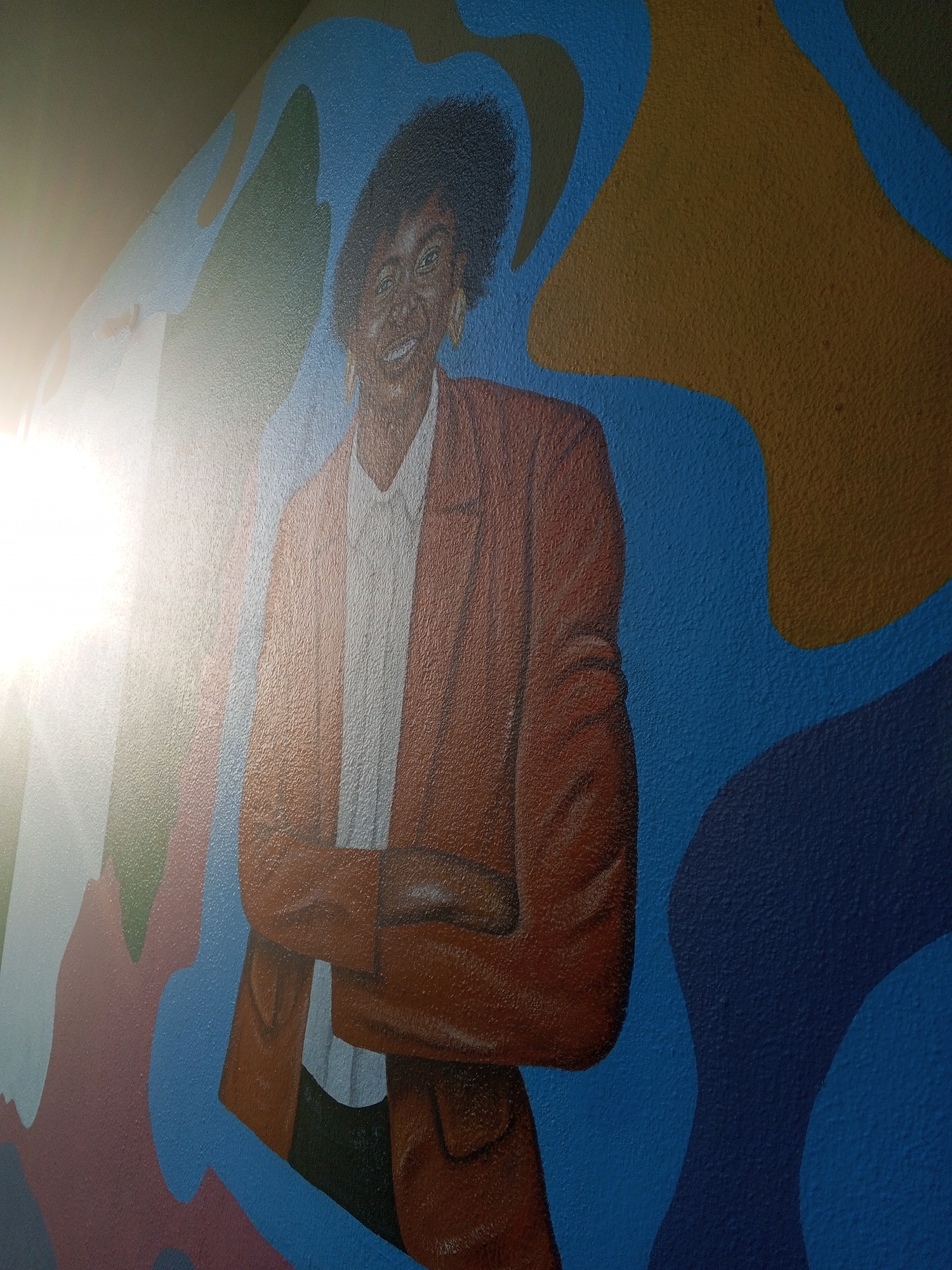 A mural depicts a dark-skinned woman with short, curly hair, wearing a white collared shirt and an open rust-colored blazer, with her arms crossed. The mural is painted on a textured wall with a blue background with abstract shapes in tan, green, white, and burgundy. No information about the location in the city of Tawpo, Nigeria, is available.