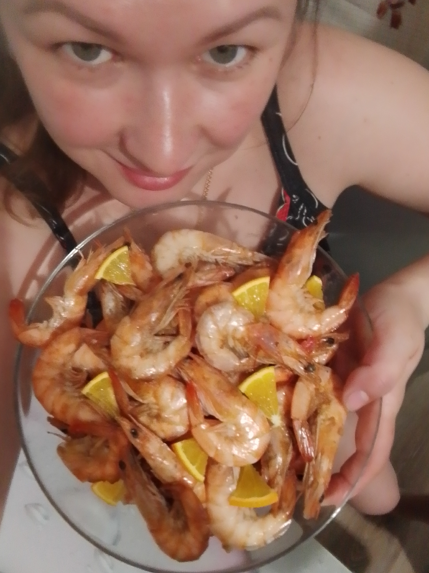 A woman is holding a glass bowl filled with cooked shrimp and slices of orange. The woman has fair skin, green eyes, and is wearing a dark top with a floral pattern. The shrimp appear to be cooked and seasoned. The background is somewhat indistinct, but the location is Noviy, Russia.