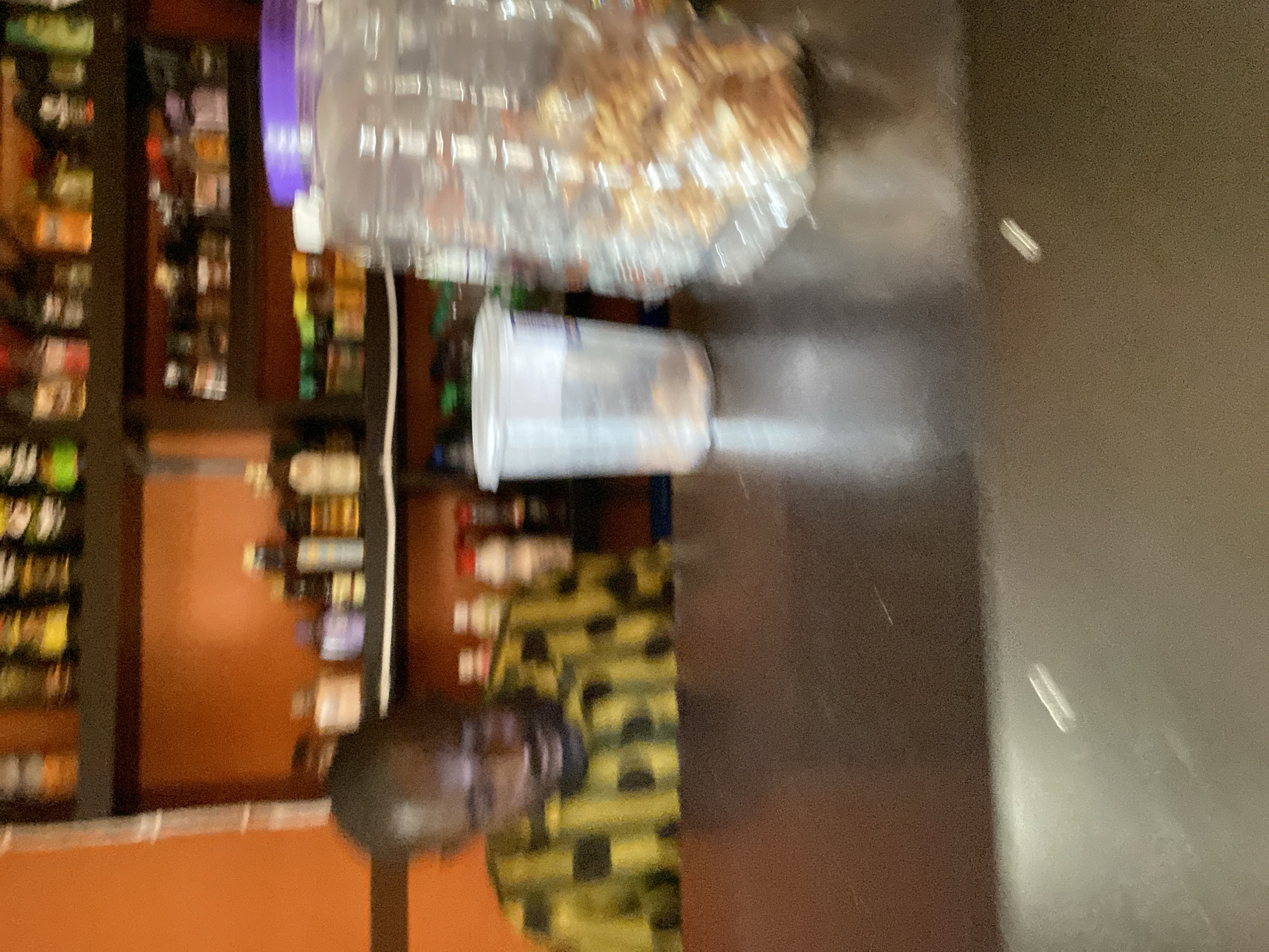 The image captures an indoor scene, likely a bar or shop, in Bukuru, Nigeria. A person, appearing to be male, is visible behind a dark countertop. He is wearing a yellow and black checkered shirt and is partially obscured by the countertop. The background shows shelves stocked with various bottles, suggesting alcoholic beverages or other retail items. On the countertop, there is a clear plastic cup with a white lid and a large clear plastic container filled with what appears to be snacks, possibly peanuts or chips. The lighting in the scene is somewhat dim, and the image is slightly out of focus, contributing to a blurry overall appearance. The walls in the background are painted orange.