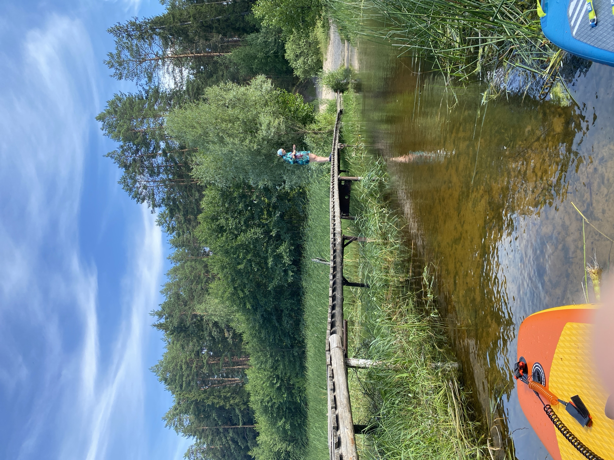 The image depicts a serene natural scene in Yal'chik, Russia. In the foreground, the edge of an orange and yellow paddleboard is visible in the shallow, clear water. The water reflects the sky and surrounding trees, showing a sandy bottom in places. A narrow, weathered wooden bridge crosses the water, extending from the lush green grassy bank on the left to a sandy path on the right.
A person wearing a blue and green patterned t-shirt and shorts stands on the bridge, looking down at the water. They are wearing a light-colored bandana on their head. Tall green reeds and grasses line the edges of the water and the bridge. In the background, a dense forest of tall pine and deciduous trees forms a natural, verdant backdrop under a bright blue sky with wispy white clouds. The overall atmosphere is tranquil and suggests a warm, sunny day.