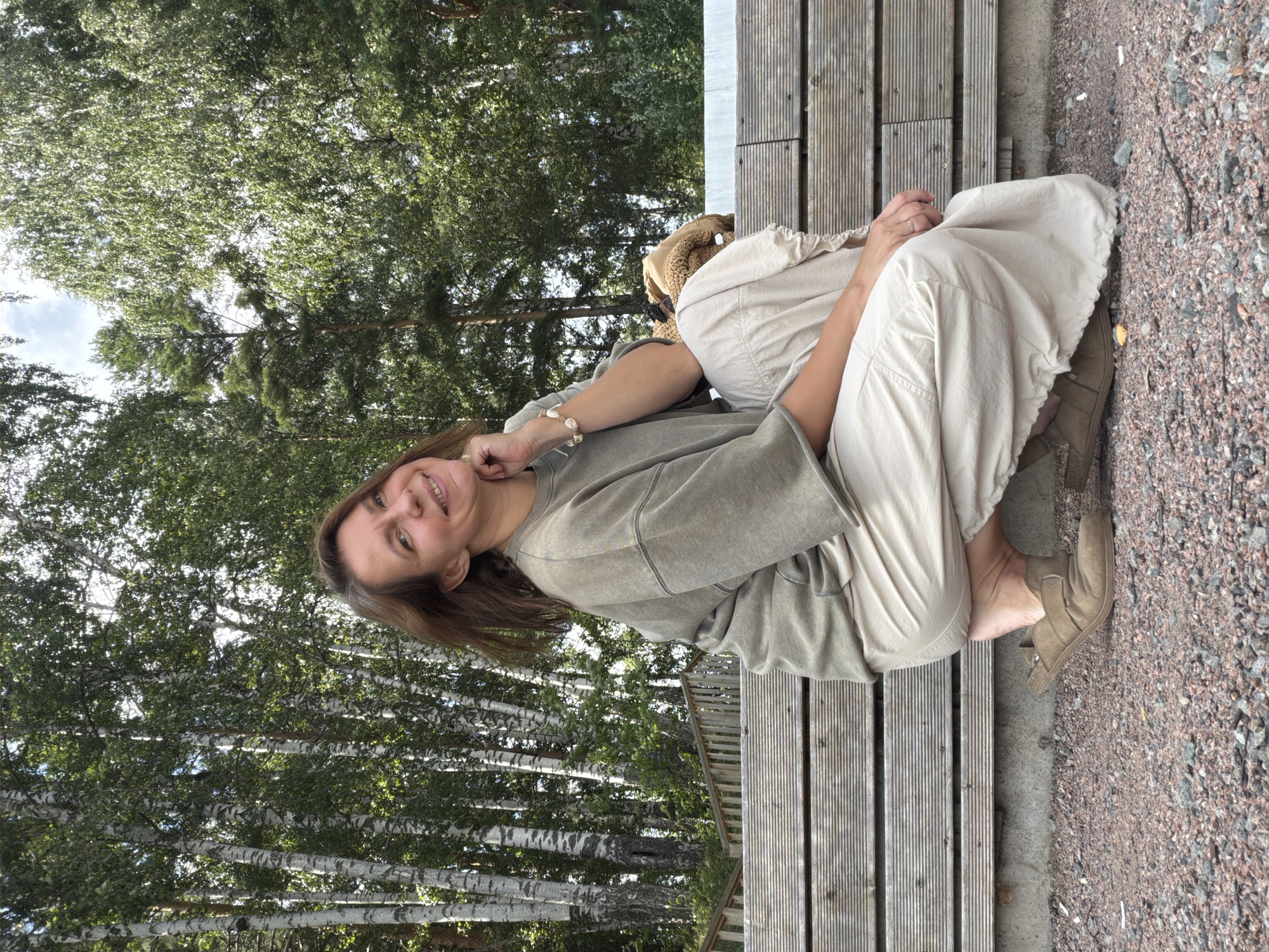 A woman with brown hair and fair skin kneels on gravel in front of a wooden bench. She is wearing an oversized olive-green sweatshirt and loose, cream-colored pants. Her right hand is resting on her chin, and she is smiling at the camera. Behind her, a backdrop of birch trees and other greenery is visible, suggesting an outdoor, possibly park-like setting. The lighting indicates it is daytime. The location is stated to be the city of Chaikovsky, Russia, implying this is likely a public or recreational area within or near the city.