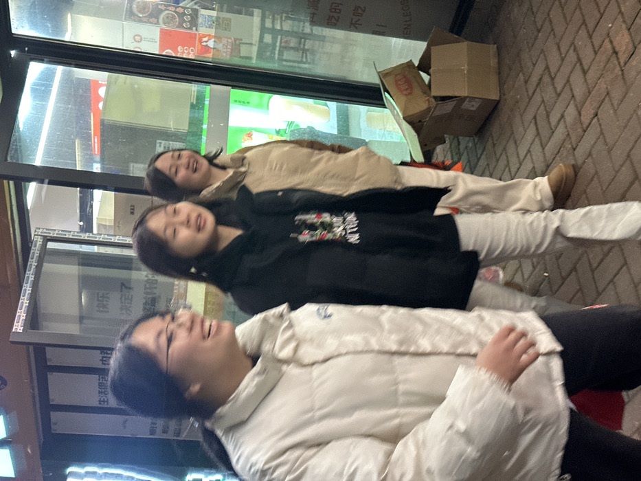 The image depicts an outdoor, nighttime scene on a brick-paved sidewalk in Jiangjin, China, adjacent to commercial storefronts. Three East Asian females are present, arranged linearly from left to right. The leftmost individual, wearing eyeglasses, a light-colored puffer jacket, and dark pants, has her mouth open in a laugh and is looking to her left. The middle individual, wearing a black hooded sweatshirt with a visible graphic design and light-colored pants, has a slight smile. The rightmost individual, wearing a beige puffer jacket and light-colored pants, displays a wide laugh. All three appear to be engaged in social interaction. In the background, illuminated storefronts with large glass panels are visible, displaying Chinese characters such as "生活" (life), "快乐" (happy), "决定了" (decided), and other characters related to food, including a partial "EN LEGS" text. Two brown cardboard boxes, one open and partially collapsed with a smaller box on top, rest on the sidewalk to the right of the individuals. Artificial lighting from the stores illuminates the immediate area.