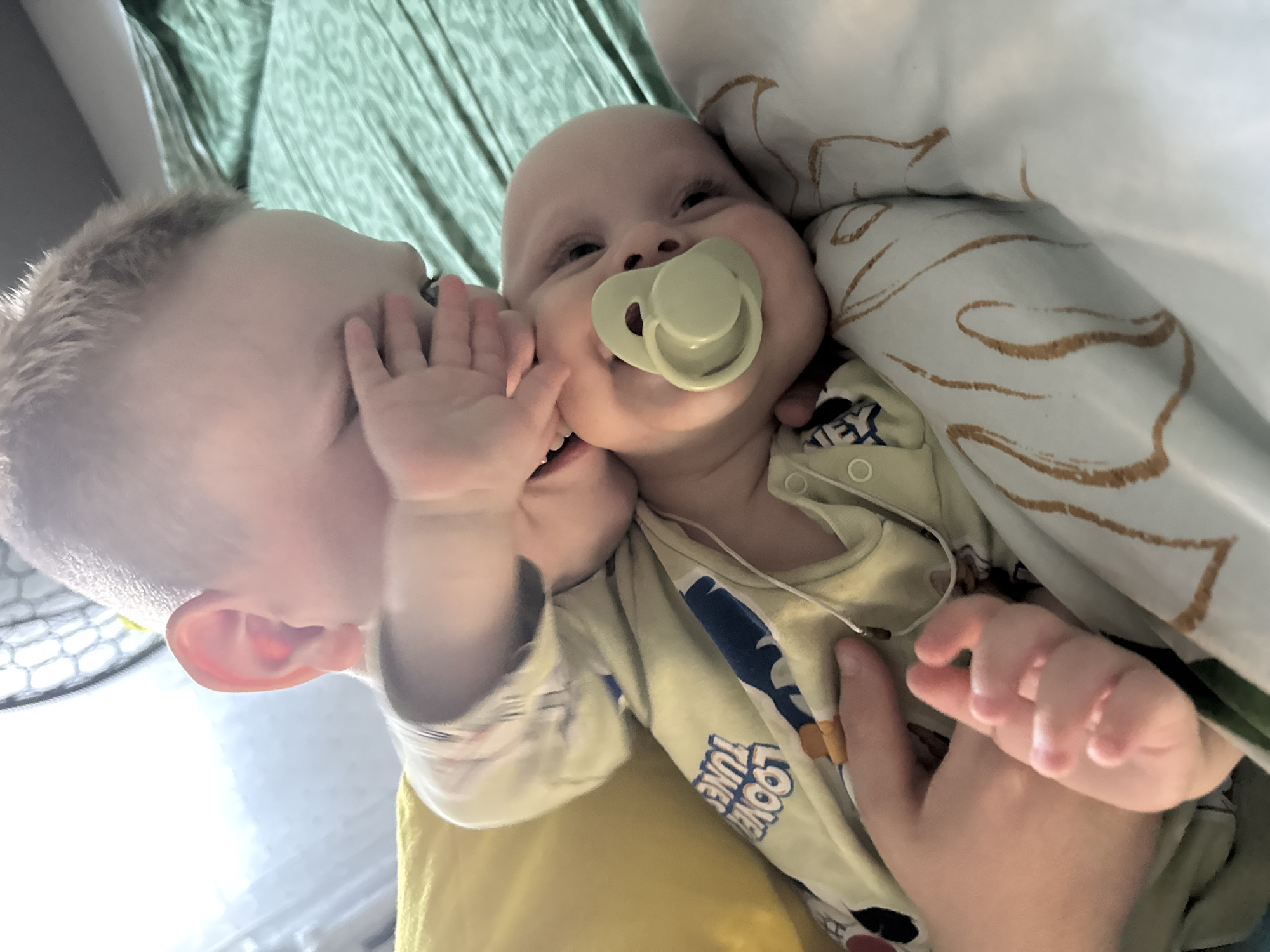 Two young children are captured in a close-up, tilted shot, likely on a bed or sofa. On the left, an older child with light hair is leaning in towards an infant, their left hand gently touching the infant's cheek or chin. The older child's expression is partially obscured, but their posture suggests playful interaction, possibly whispering or making a sound to the baby. The infant, positioned towards the right, looks up and slightly to the left, appearing content with a pale green pacifier in their mouth.
The infant is wearing a light-colored onesie that features the visible text "LOONEY TUNE" as part of a graphic pattern. The older child wears a light-colored top, possibly yellow or light green. The background includes a light green patterned fabric on the left and a white or off-white blanket with abstract brown squiggle patterns on the right. The soft, diffuse lighting indicates an indoor setting during the daytime. The image captures a sweet and affectionate moment between the siblings. This scene takes place in Chaikovsky, Russia.